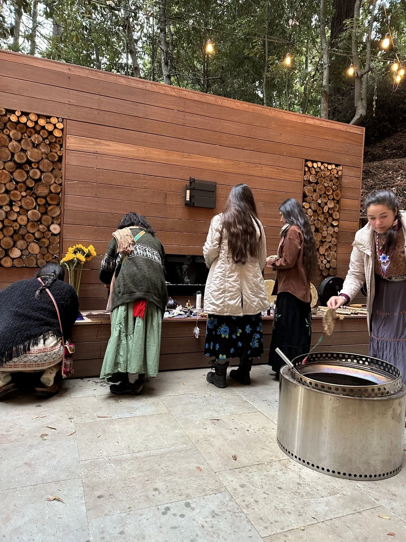 A group of women gather around a wooden table outdoors, with firewood stacked behind them and string lights hanging above, in a wooded area with trees.