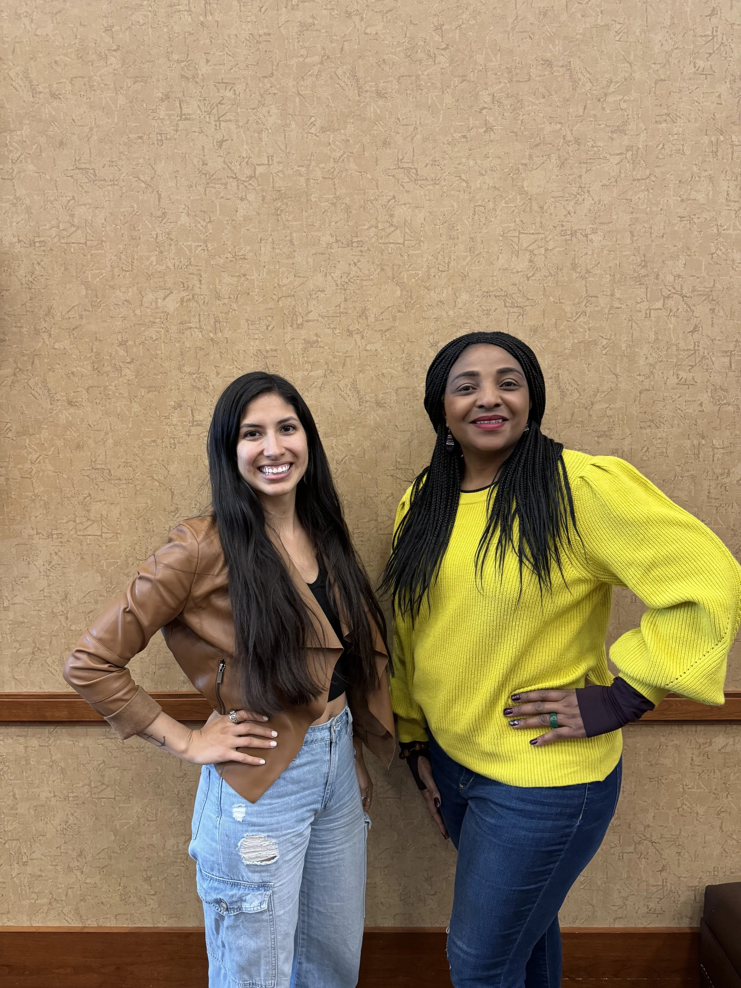 Two women standing side by side, smiling, in front of a beige textured wall. The woman on the left has long dark hair, a brown leather jacket, a black top, and light blue ripped jeans. The woman on the right has long braided hair, a yellow sweater, dark blue jeans, and colorful rings.