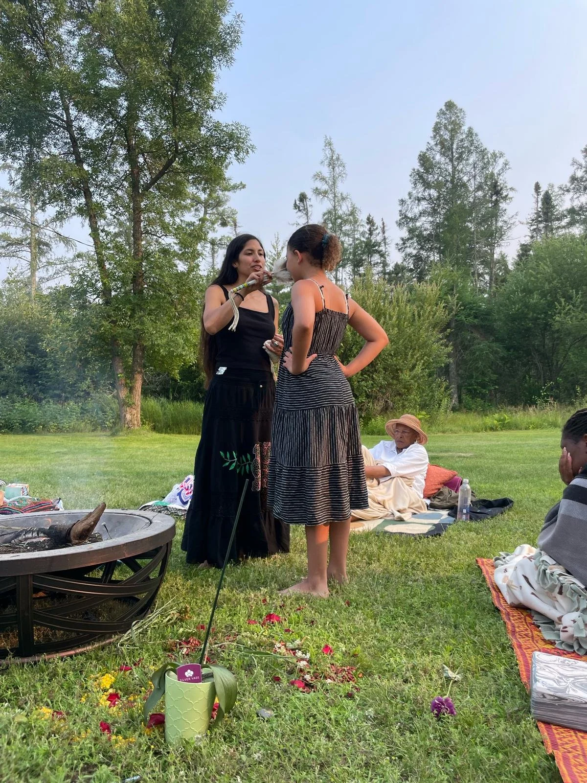 Two women standing outside on grass while talking, one with long dark hair wearing a black dress, the other with curly hair in a striped dress; several people sitting on blankets and a fire pit in the foreground, trees in the background