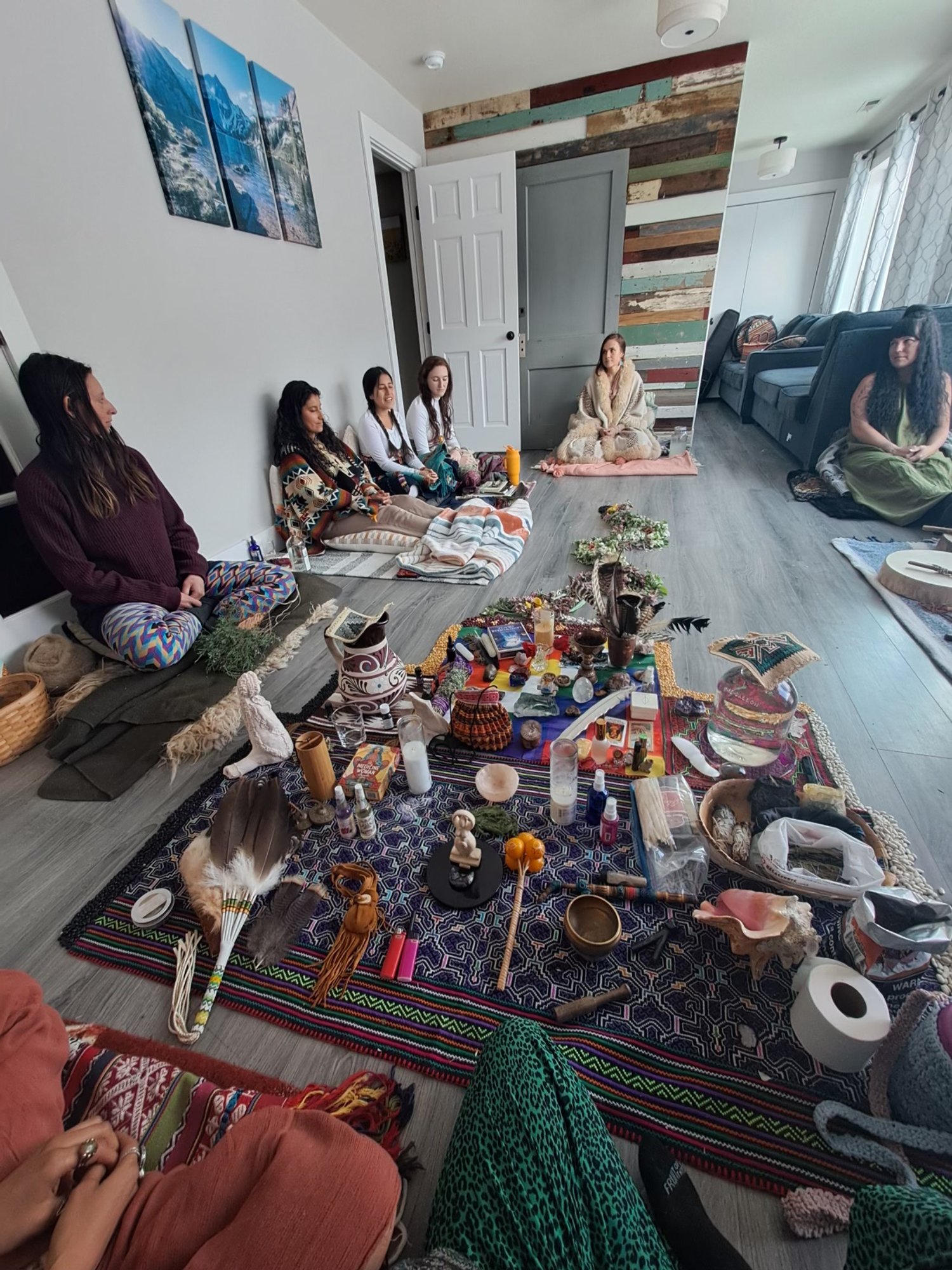 Group of women seated on the floor during a spiritual or ceremonial gathering, with a decorated altar in the center, in a cozy room with wooden wall accents and natural light.