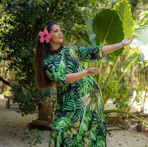 A woman in a tropical dress with huge green leaves patterns, wearing a pink flower in her hair, standing outdoors among lush greenery, holding large leaves in her hands.