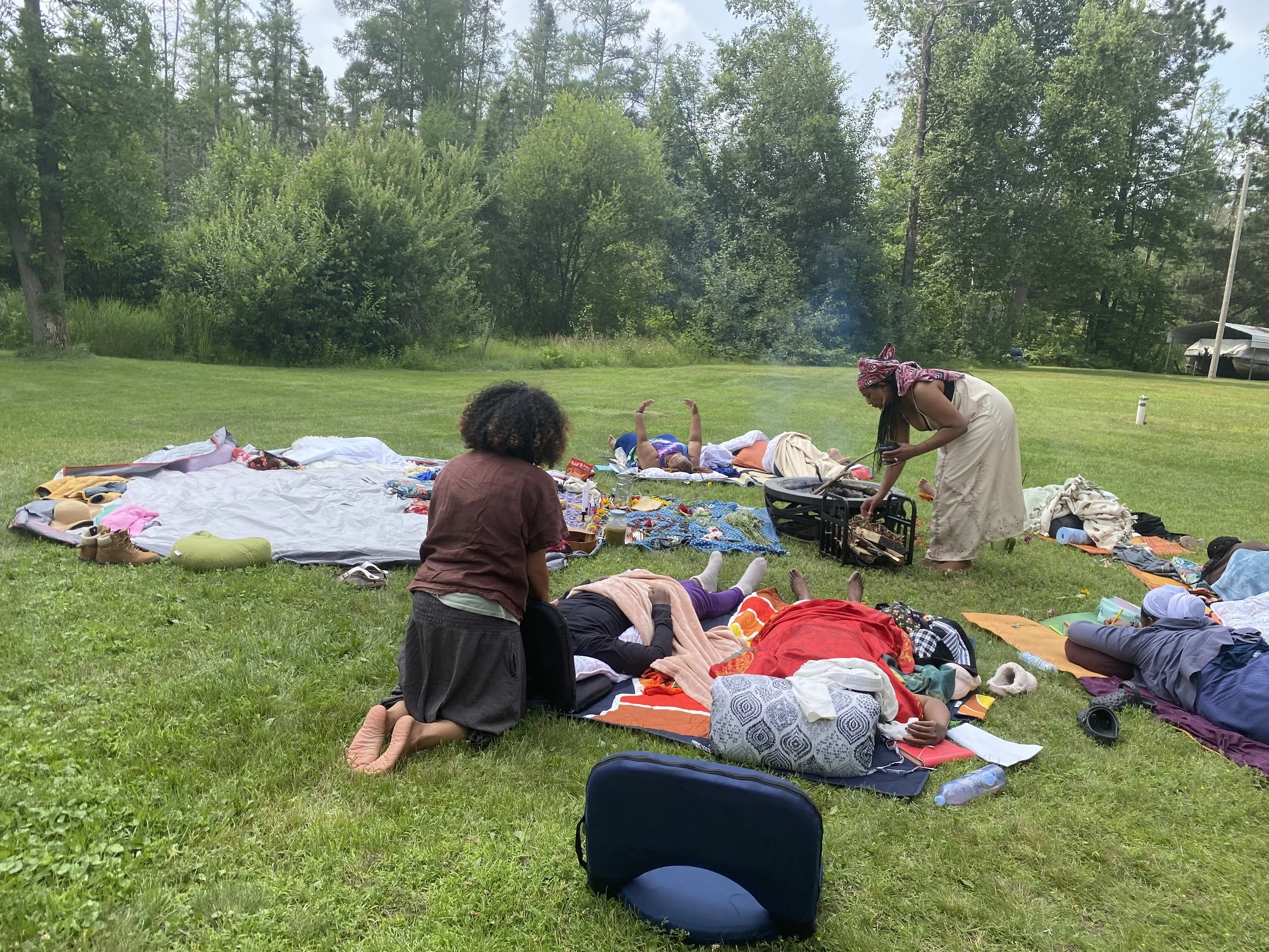Family having a picnic outdoors on a grassy field with trees in the background. Children are lying on blankets and adults are preparing food.