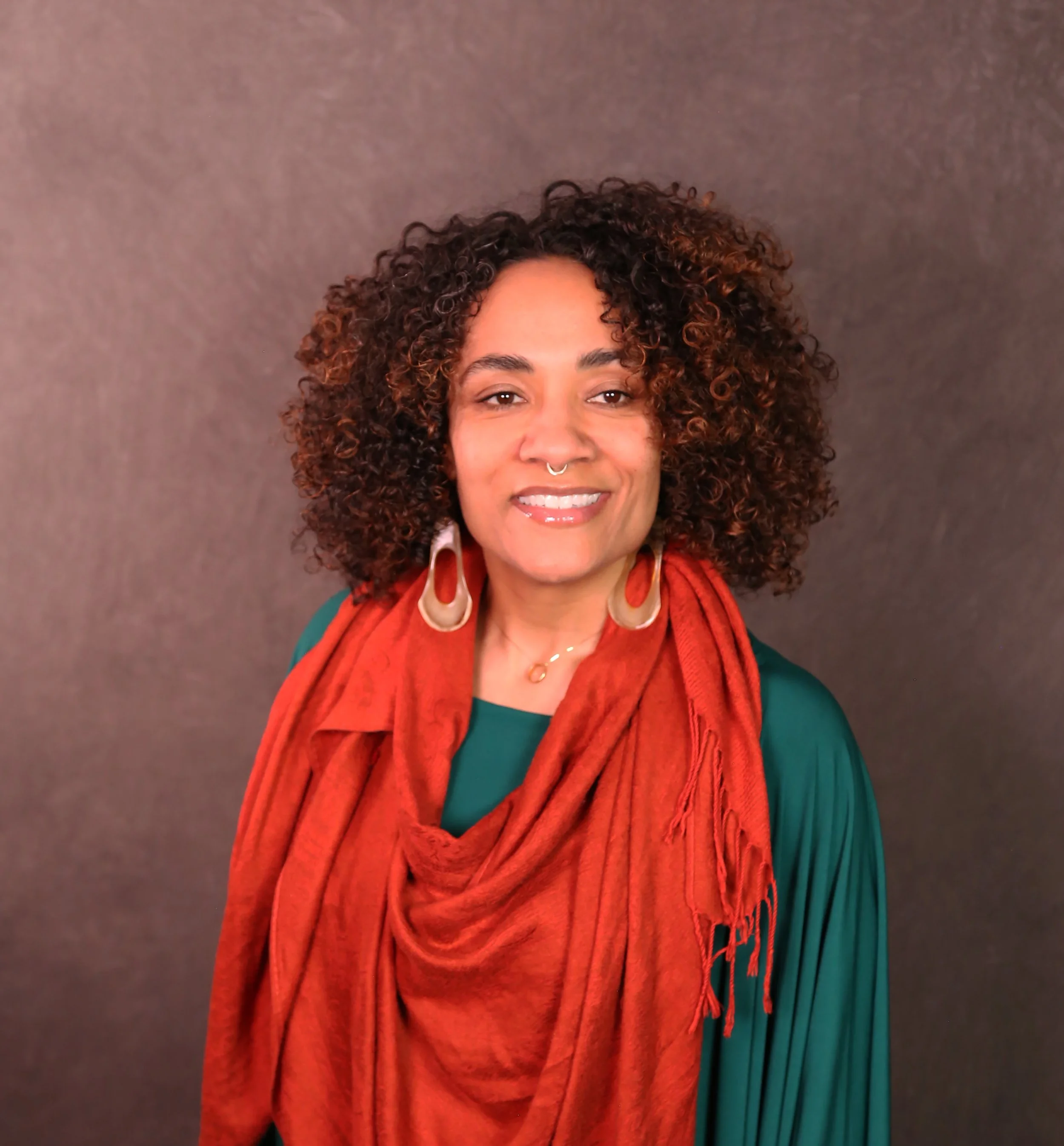 Portrait of a woman with curly hair wearing large earrings, a red scarf, and a green top against a brown background.