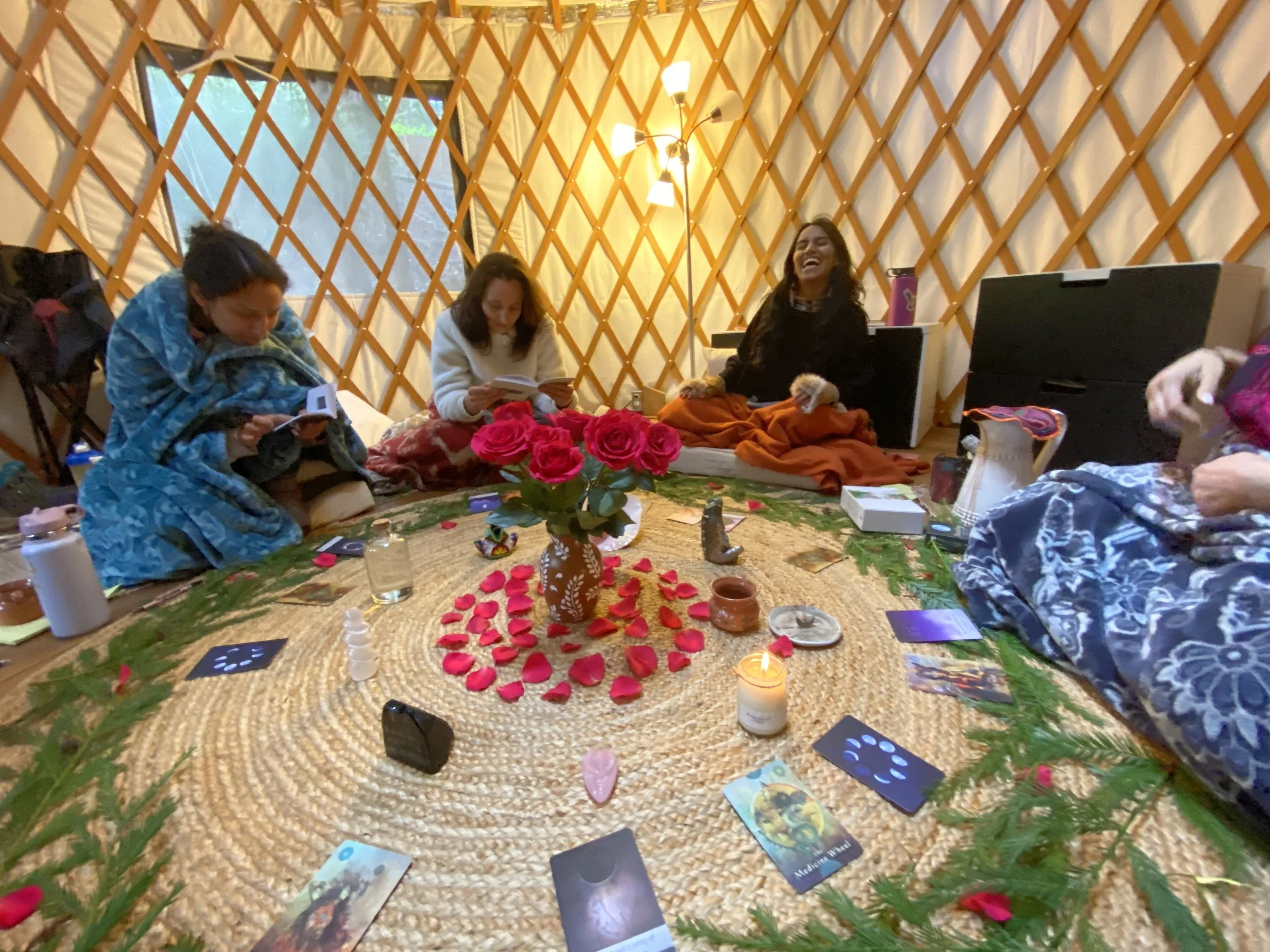 Four women sitting inside a yurt, engaged in a tarot card reading or spiritual practice, with a central bouquet of pink roses on a woven rug surrounded by tarot cards, candles, and decorative items.