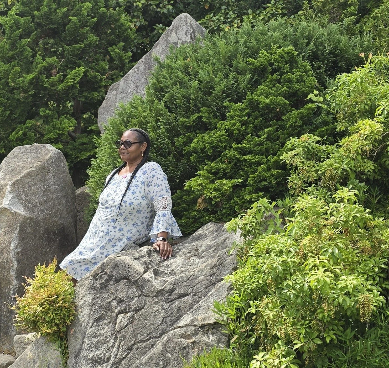 A woman in a white floral dress and sunglasses sitting on a large rock surrounded by green shrubs and trees.