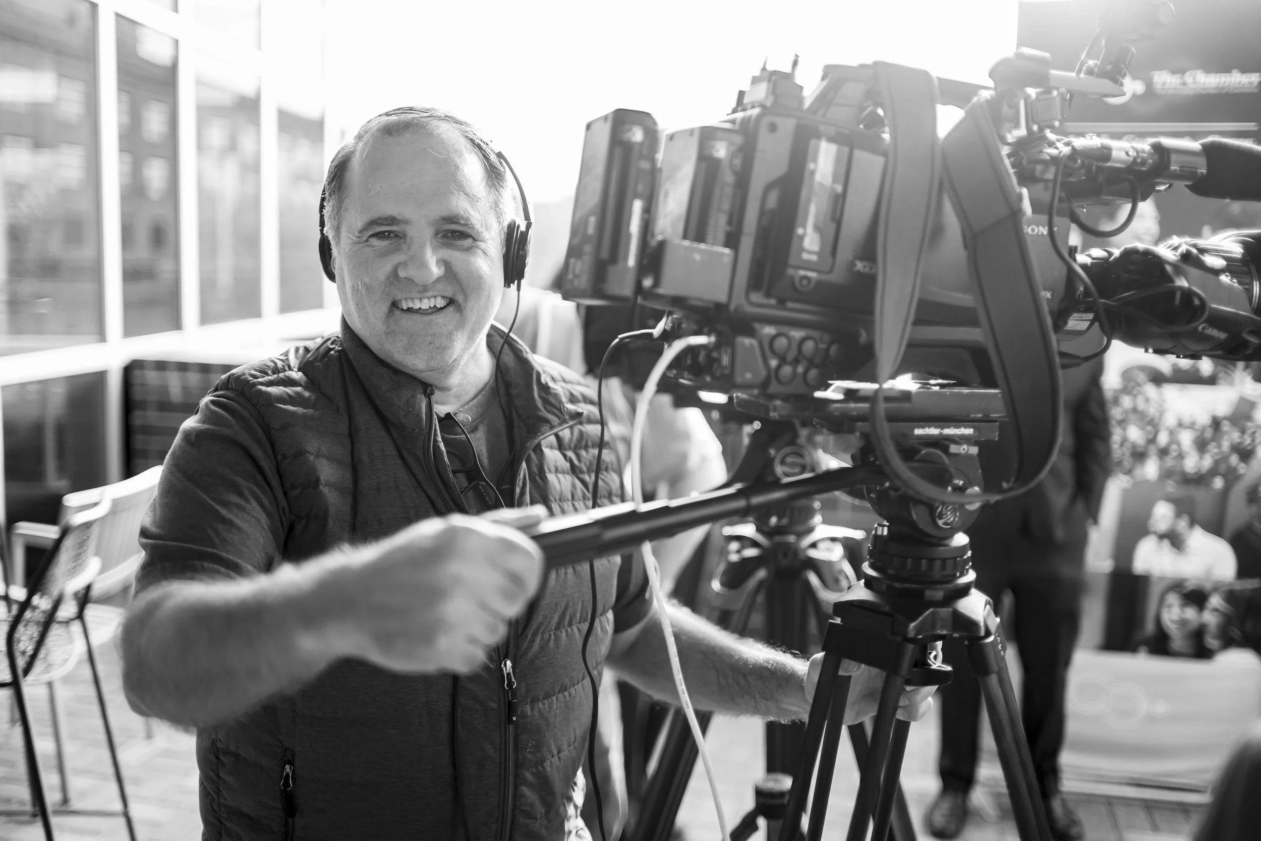A man smiling at the camera while operating a professional video camera outdoors with sunlight.