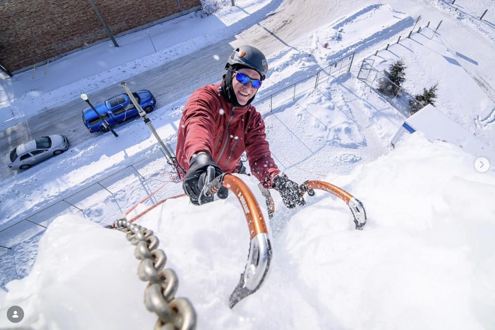 Person climbing a snow-covered wall with a safety harness, wearing a helmet and sunglasses, with parked cars and a snowy street below.