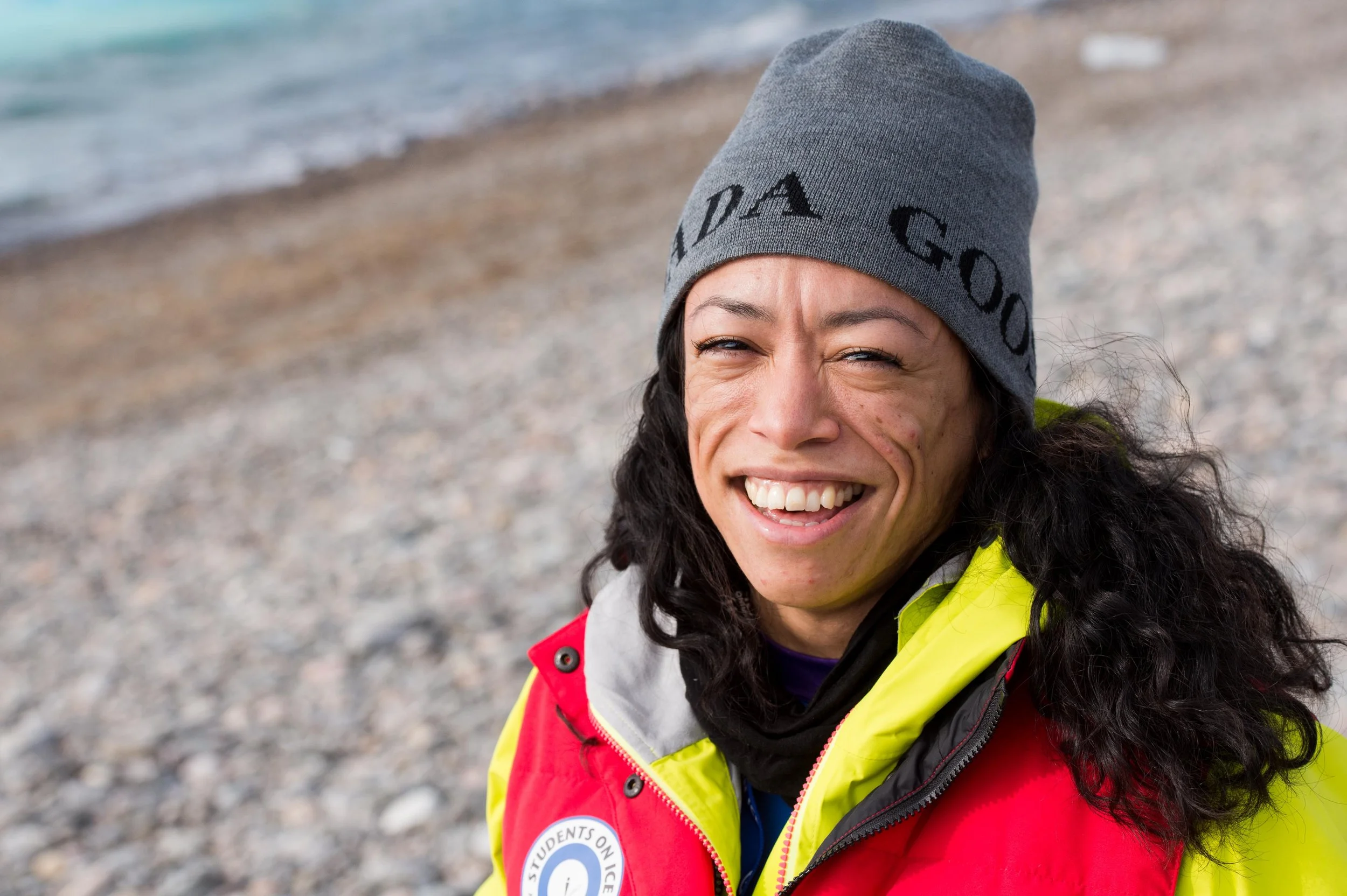 Close-up of a smiling woman with curly black hair wearing a gray beanie, a bright yellow and red jacket, and a black scarf, standing on a rocky beach with the ocean in the background.