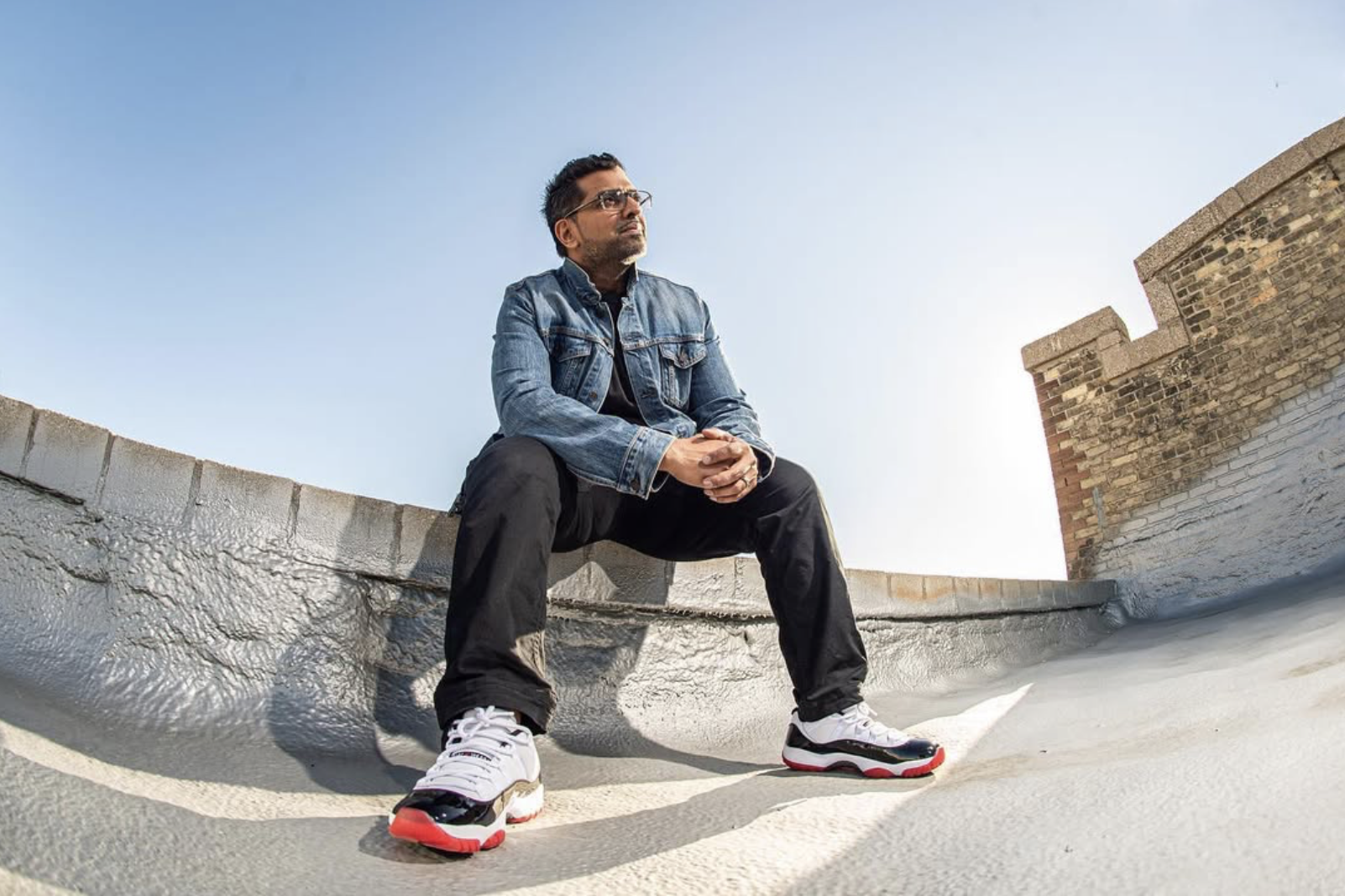 A man in a denim jacket and black pants sitting on a rooftop, with a brick wall on the right and a clear blue sky behind him.