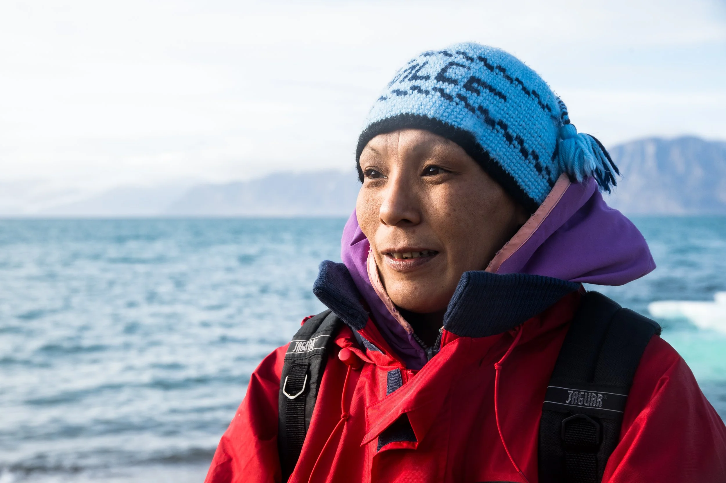 A woman wearing a colorful winter hat and red jacket standing by a body of water with mountains in the background.