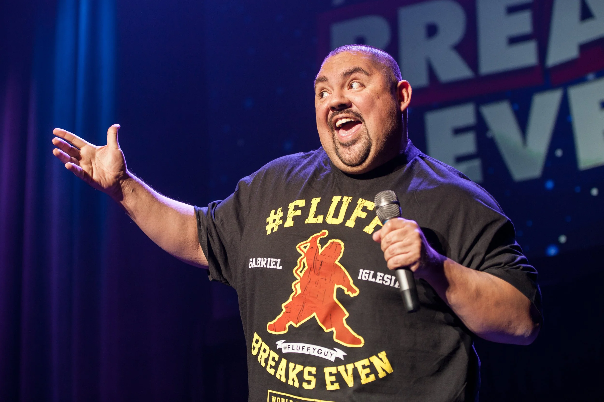 A man holding a microphone on stage, wearing a black T-shirt with #FLUFFY, a bear, and the phrase 'Fluffy Guy Breaks Even' printed on it, during a comedy show.