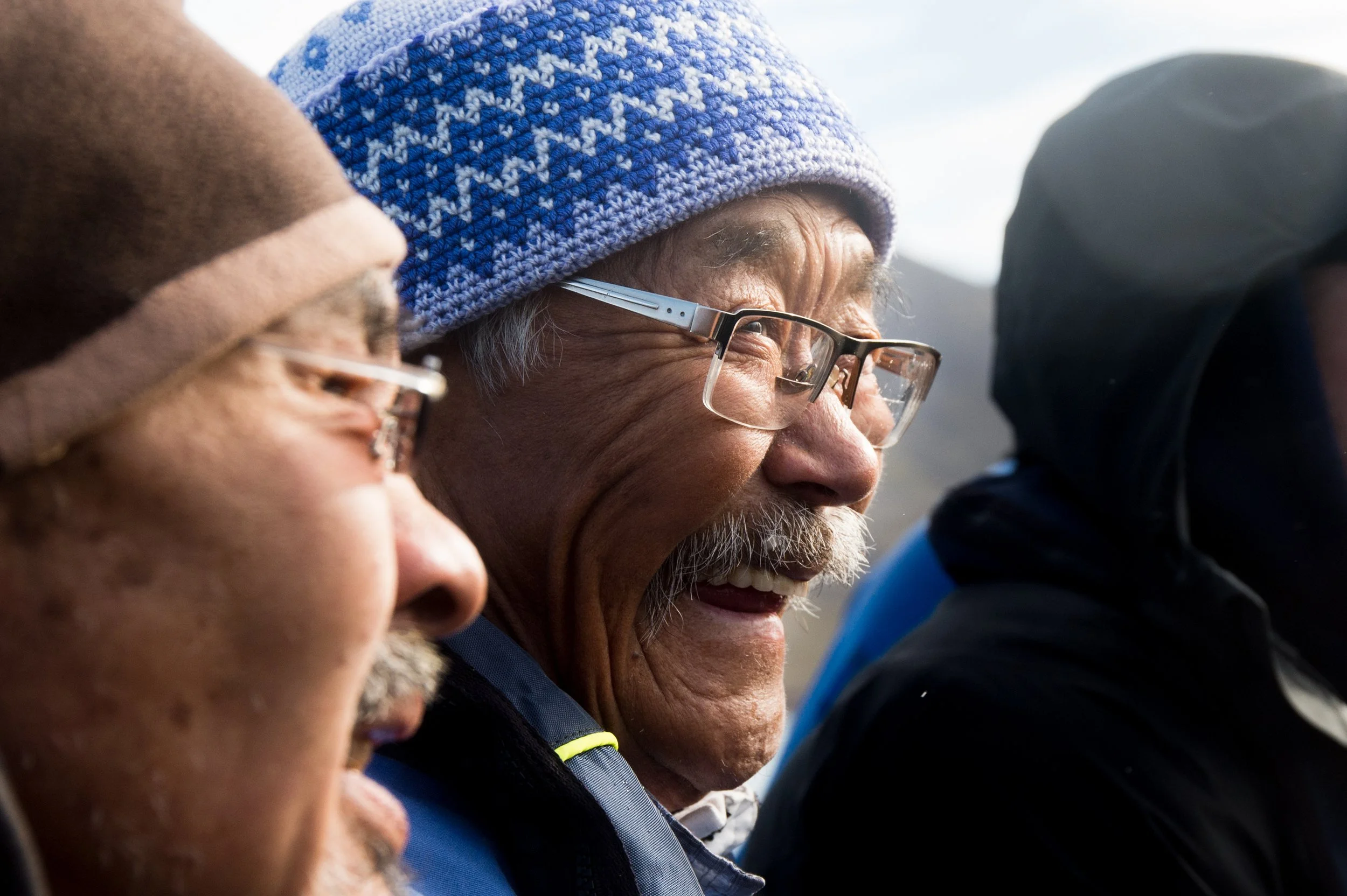 Close-up of three elderly men outdoors, smiling and wearing hats and glasses, with a mountain background.