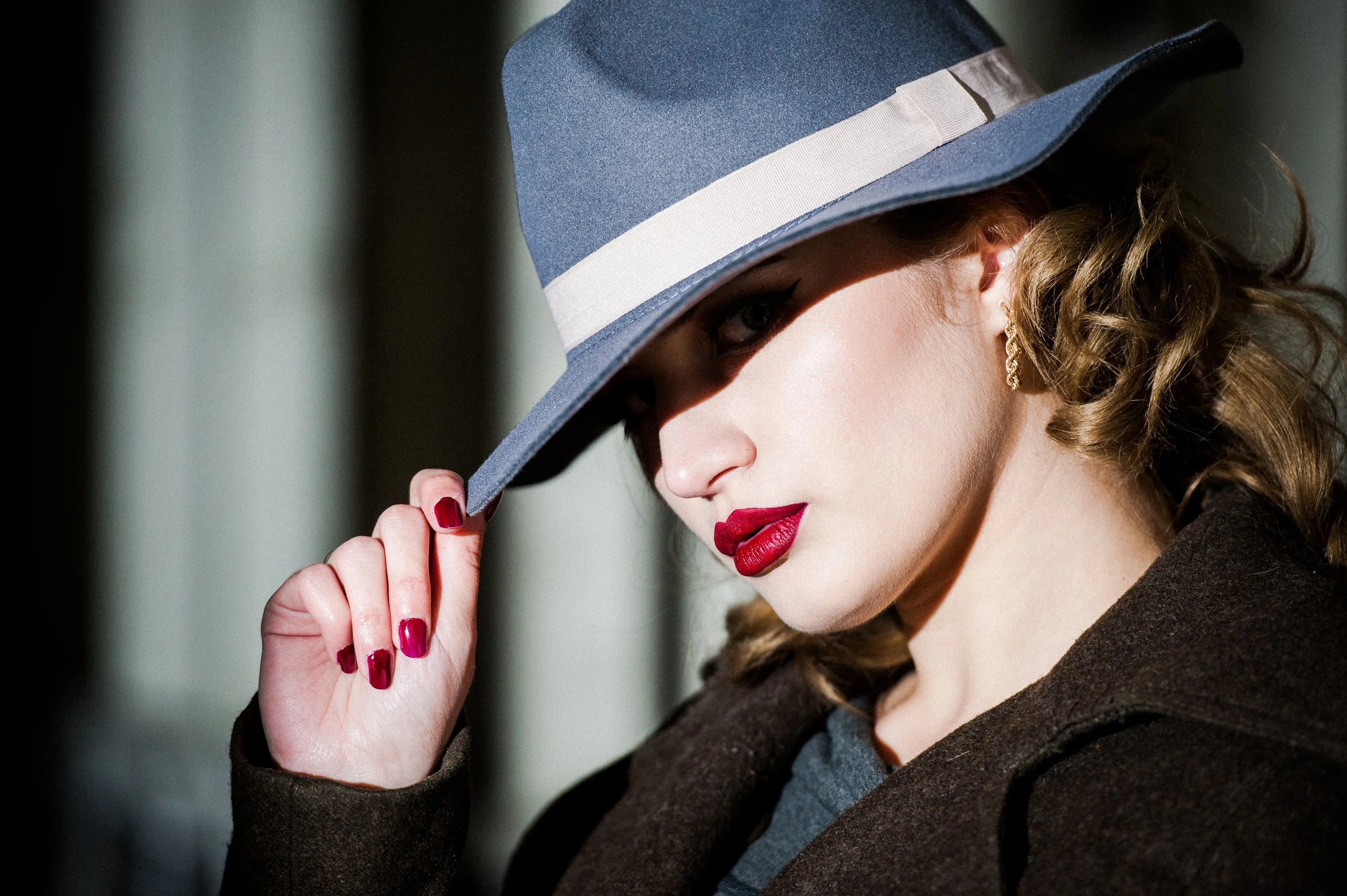 A woman with red lipstick and painted nails is wearing a blue wide-brimmed hat with a white band, turned downward to partially cover her face, and a dark brown coat.