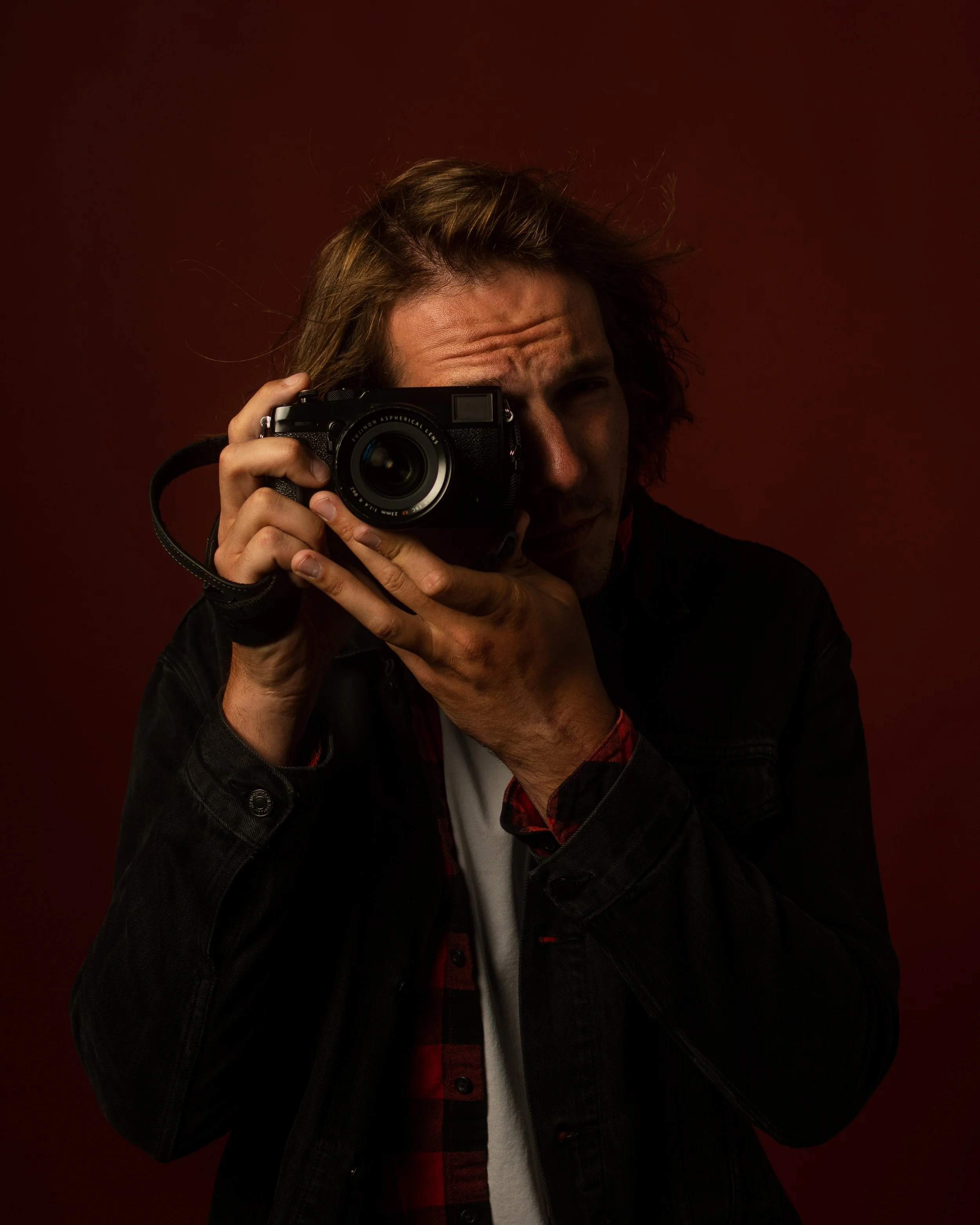A man holding a camera up to his face, looking through the viewfinder, against a dark red background.