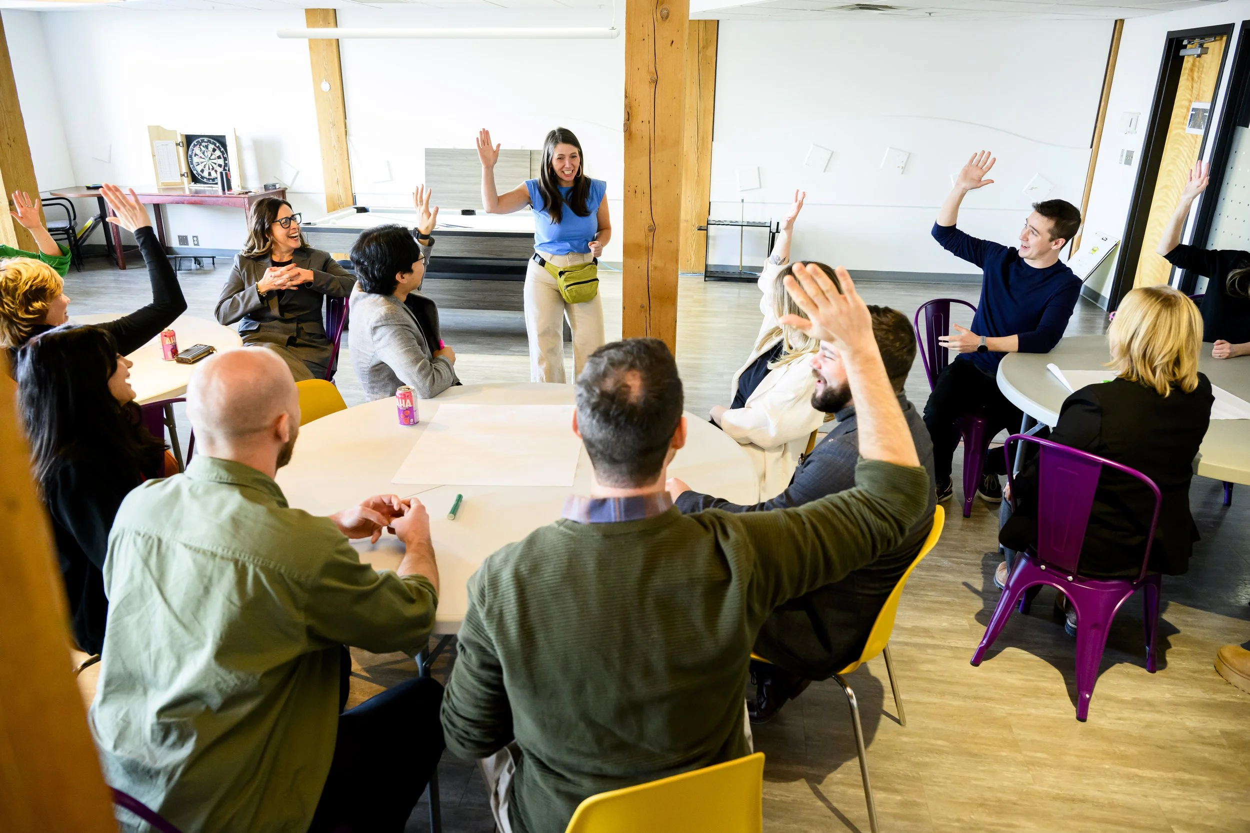 A group of people sitting around a table, raising their hands, in a room with wooden beams and a dartboard in the background.