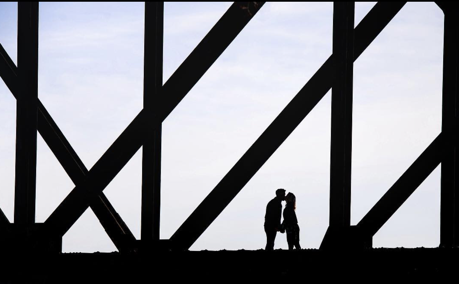 Silhouettes of a man and woman standing closely and kissing on a bridge with steel beams, against a cloudy sky.