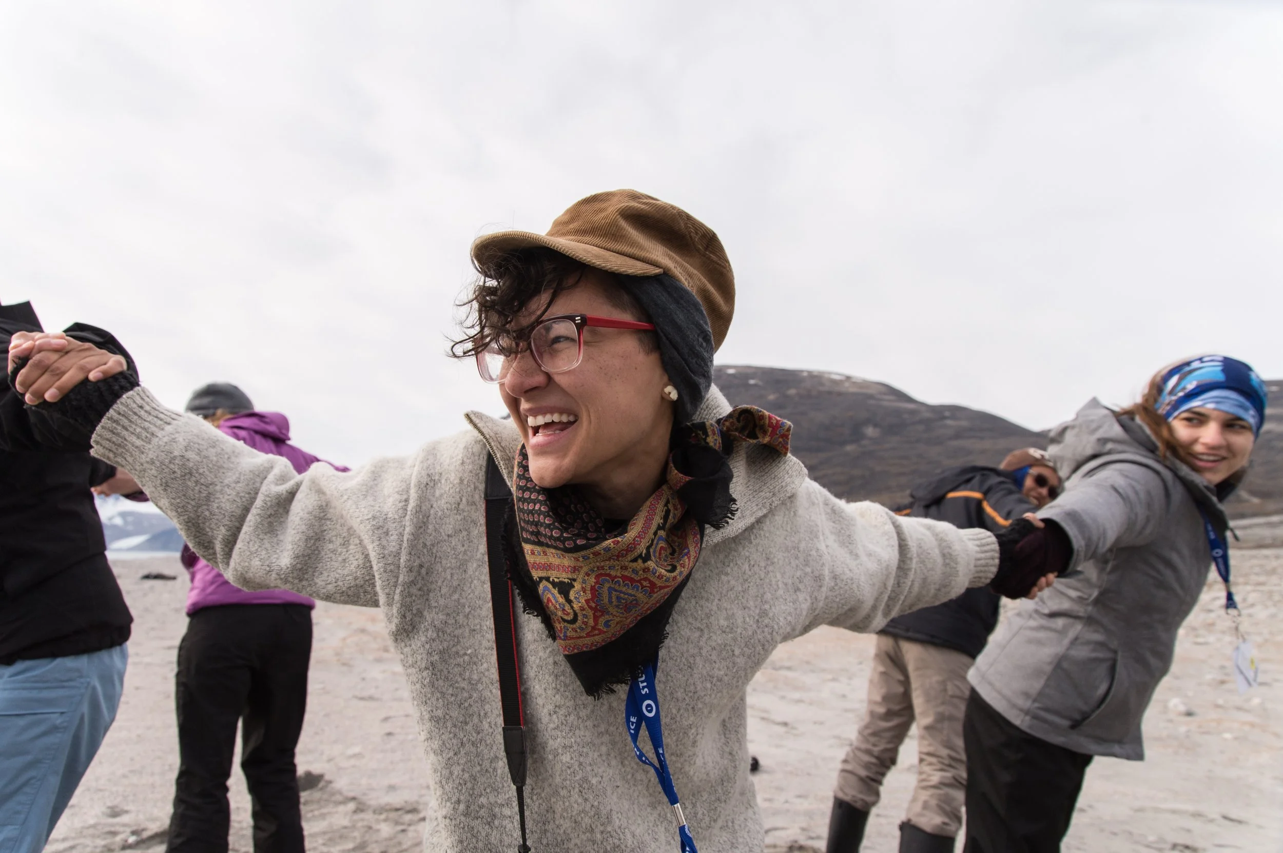 Group of people holding hands in a circle outdoors on a cloudy day, smiling and enjoying each other's company.