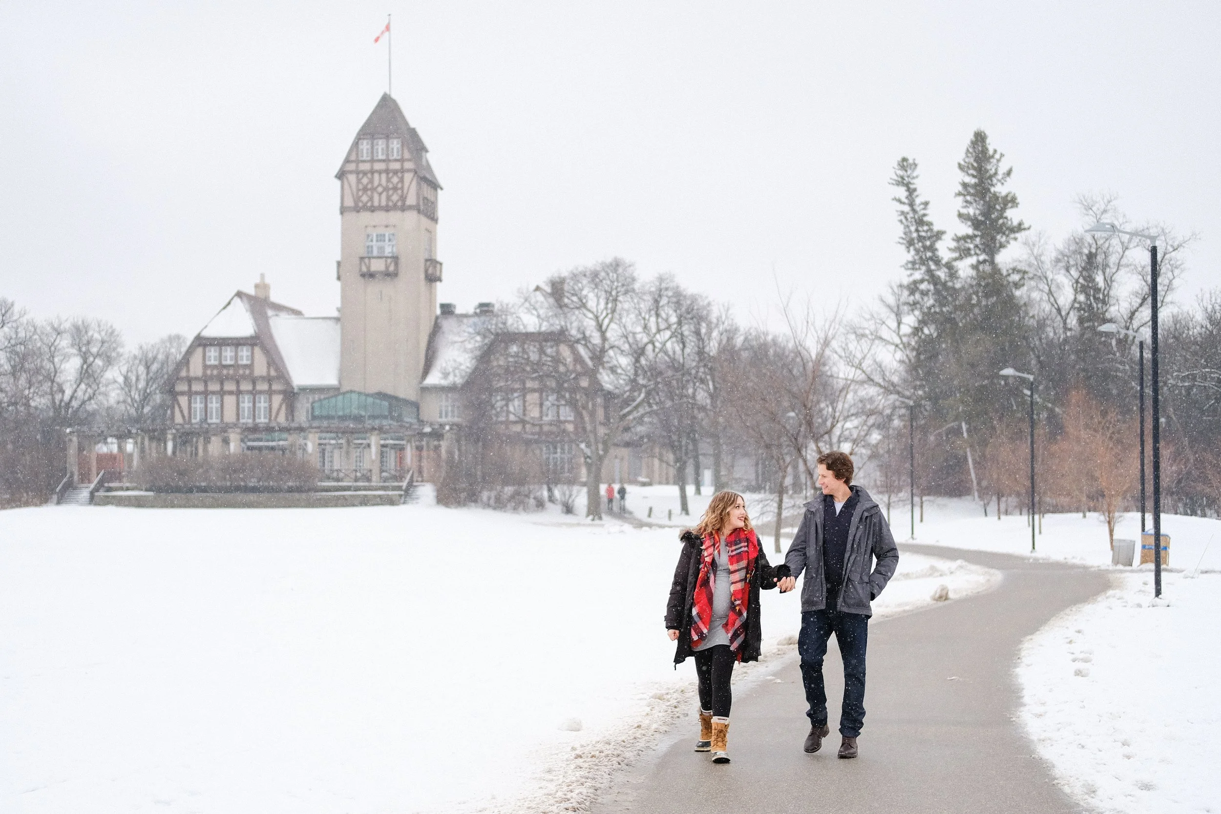 A couple walking hand in hand on a snow-covered pathway in a park, with a large, castle-like building in the background on a snowy day.