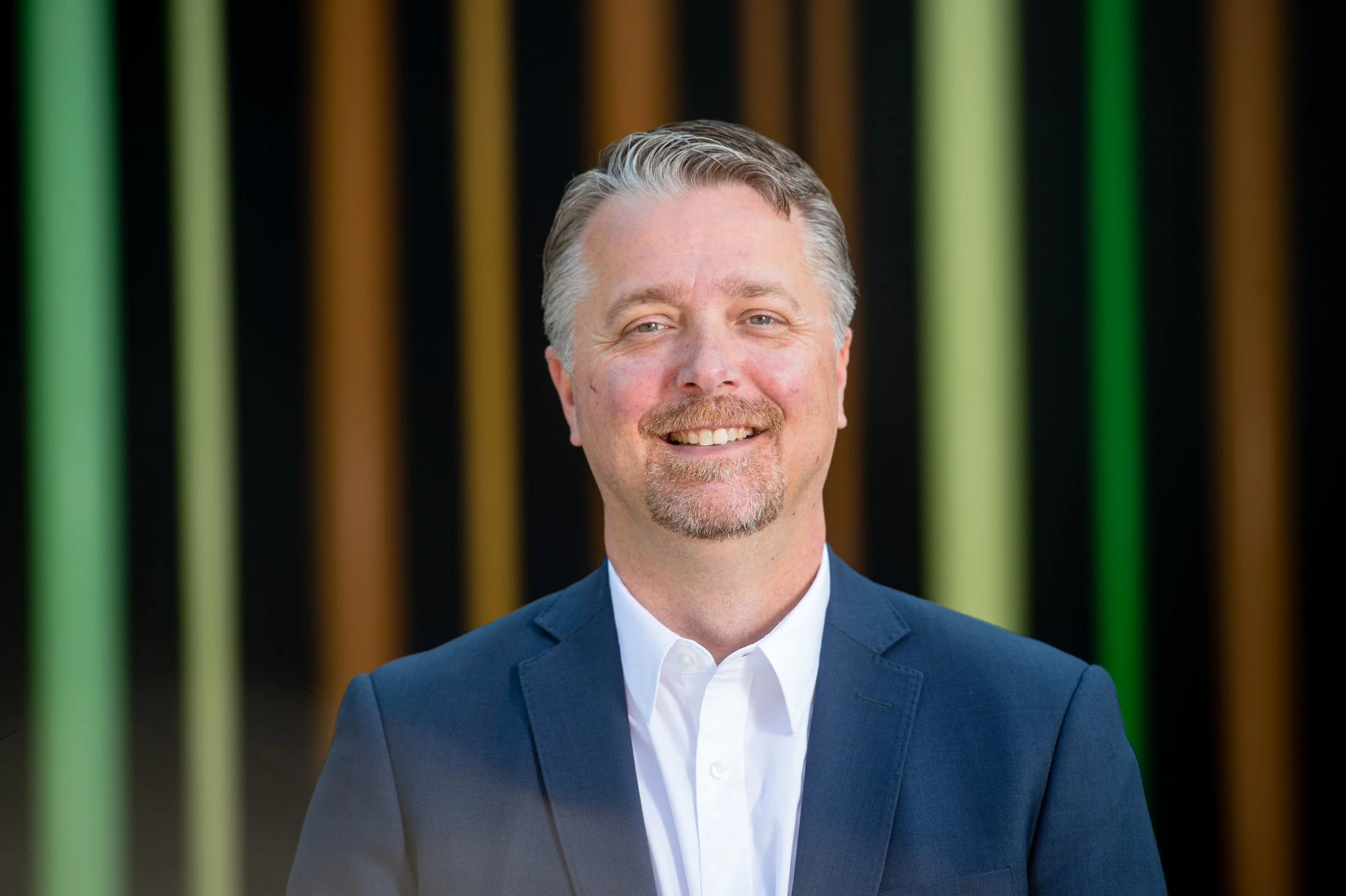A man with gray hair, a beard, wearing a blue suit jacket and a white shirt, smiling in front of a background with vertical colorful stripes.