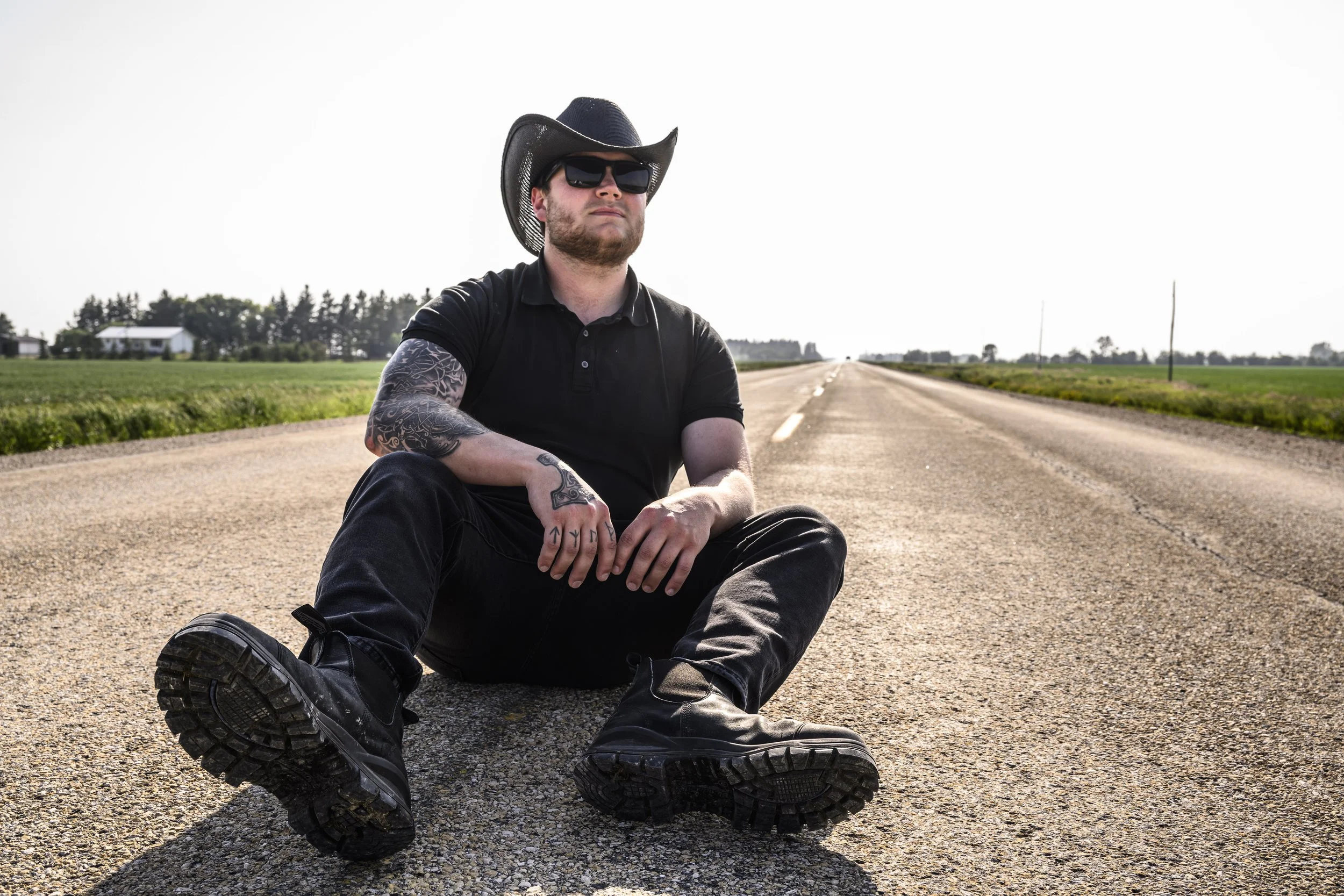 A man with tattoos, wearing sunglasses, a cowboy hat, and a black polo shirt, sitting on the road with his legs spread, against a rural landscape with green fields and houses in the background.
