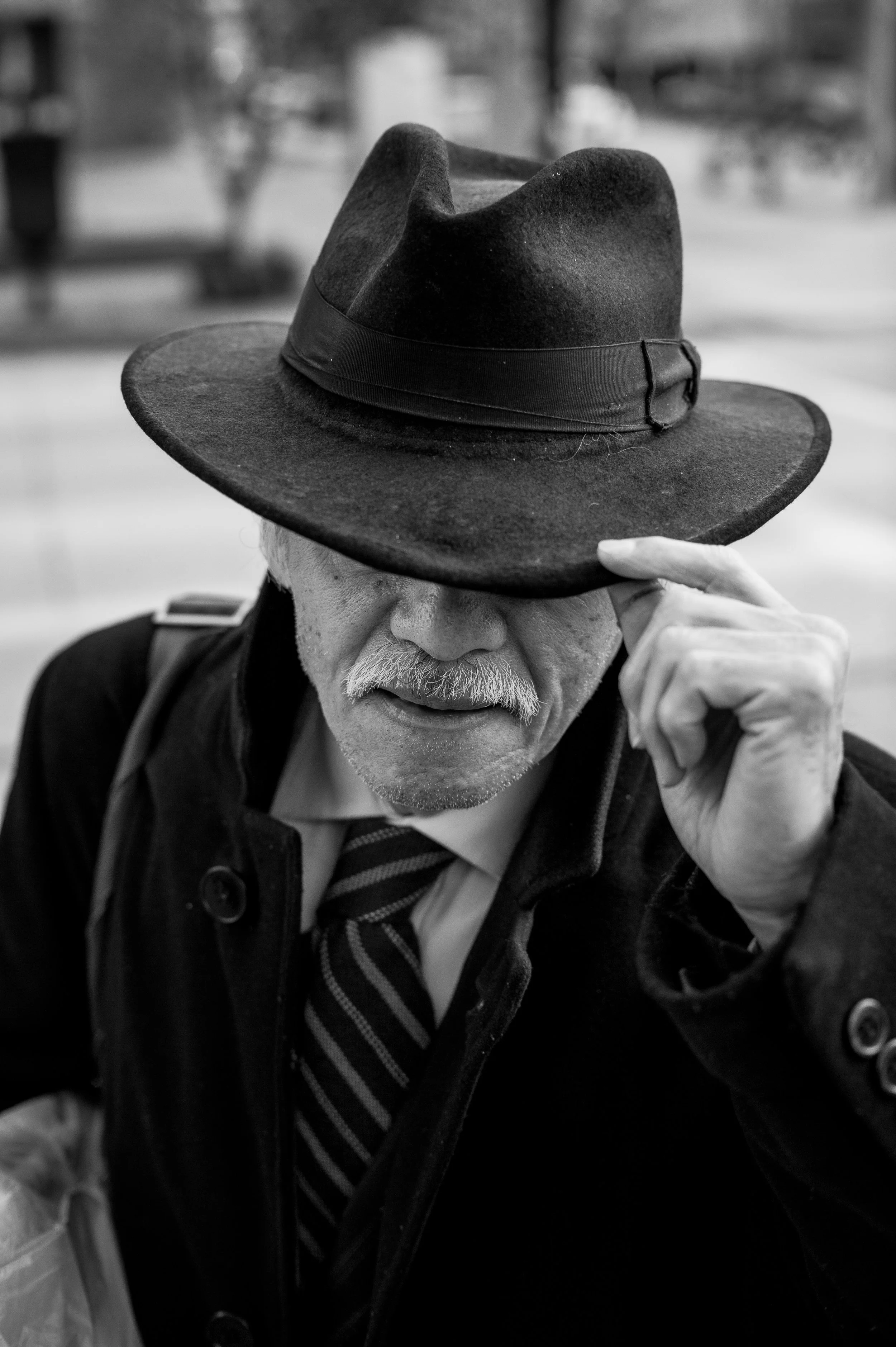A black and white photo of an elderly man with a white mustache and beard, wearing a dark coat, striped tie, and a wide-brimmed hat that covers part of his face. He is adjusting or tipping his hat with his right hand.