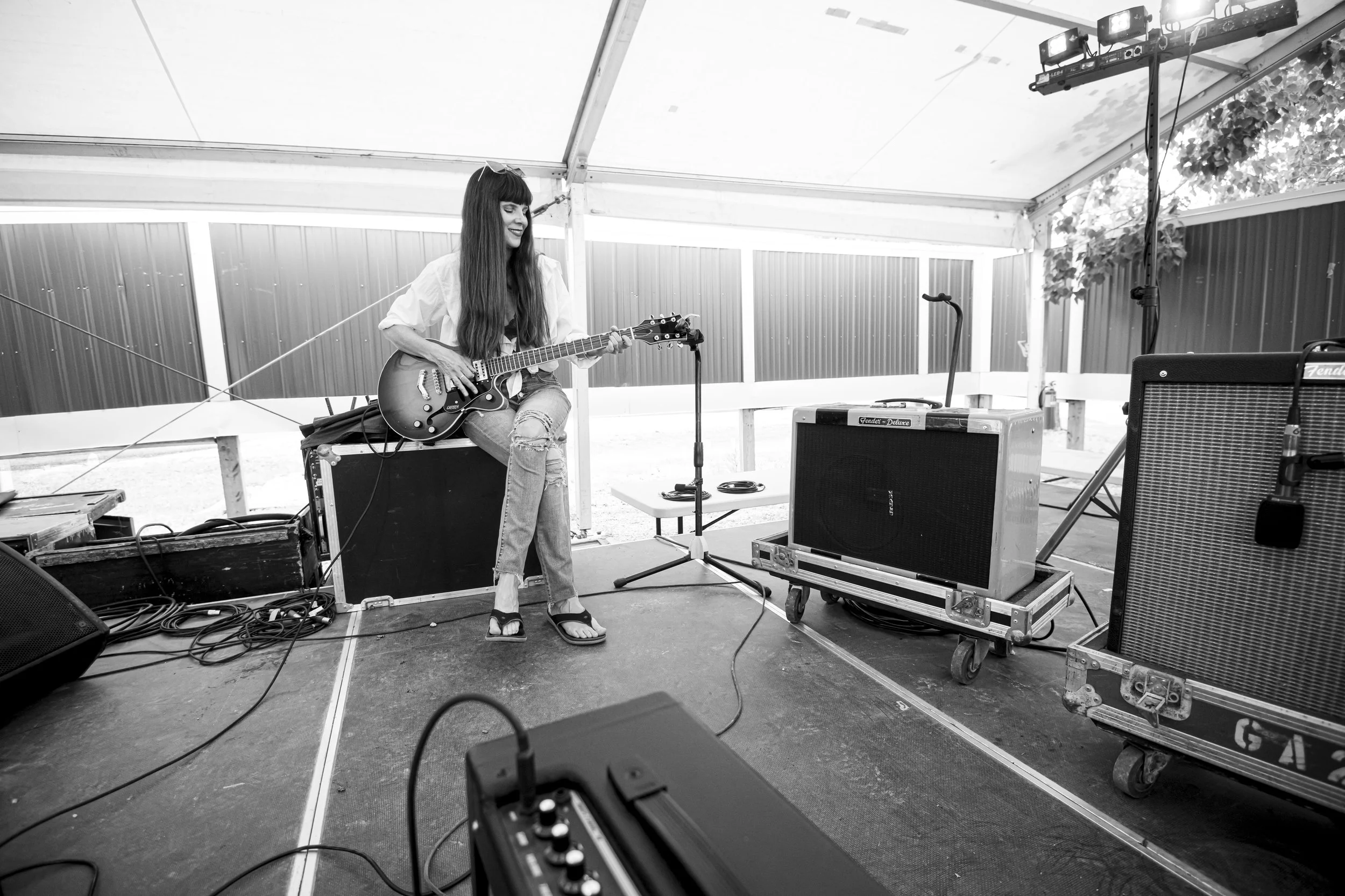 A woman with long hair playing an electric guitar on stage inside a tent with audio equipment and amplifiers around her.