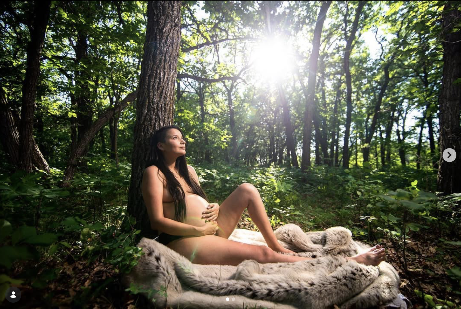 A pregnant woman sitting on a blanket in the woods during daylight, with sunlight filtering through the trees.