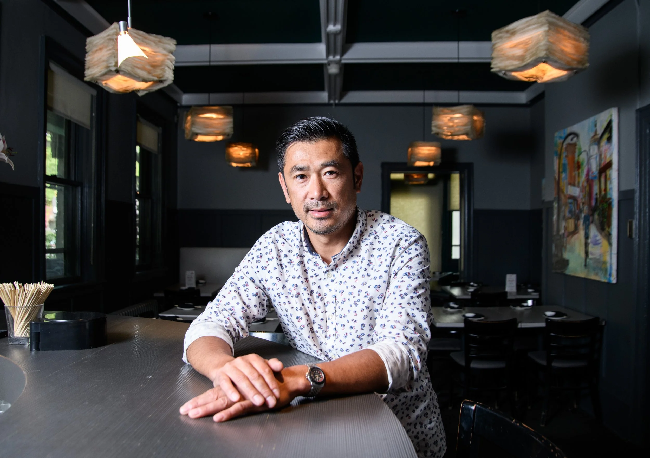 A man sitting at a restaurant bar, facing the camera, with dark walls, pendant lights, and artwork in the background.
