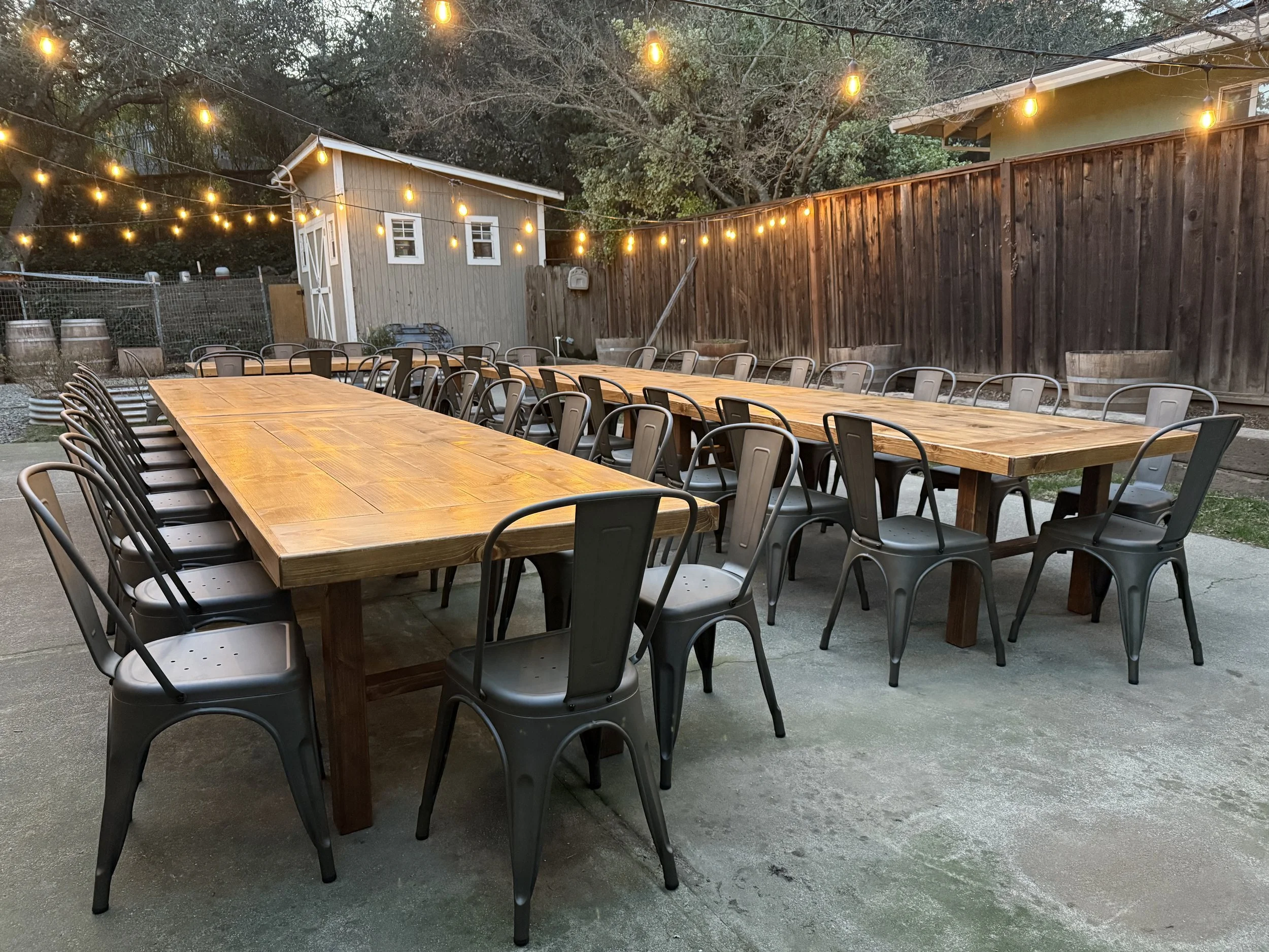 Outdoor patio with two long wooden tables and metal chairs, string lights hanging overhead, and a small shed in the background.