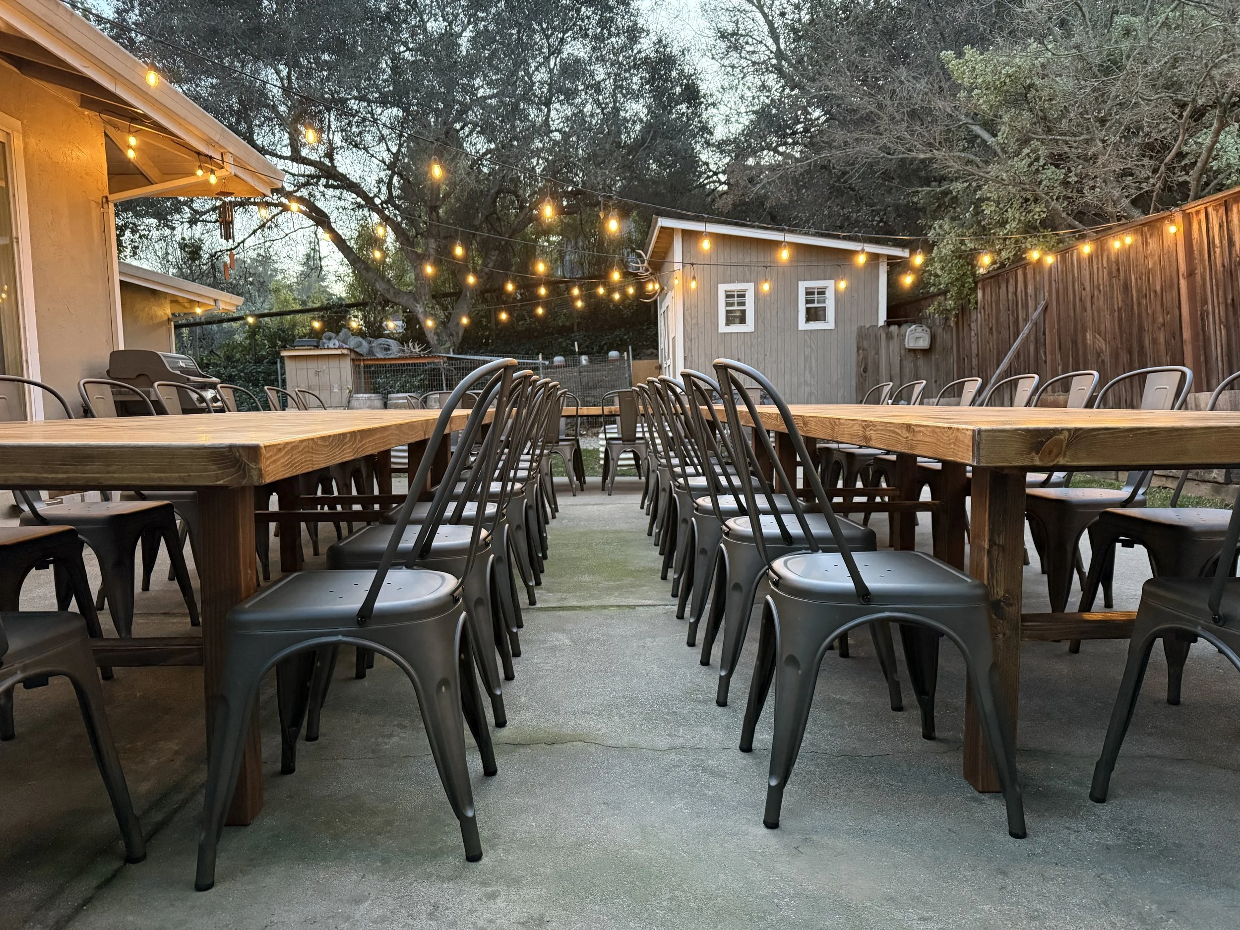 Outdoor patio with two long wooden tables and black metal chairs, string lights hanging overhead, a small shed, and a wooden fence, with trees in the background at dusk.