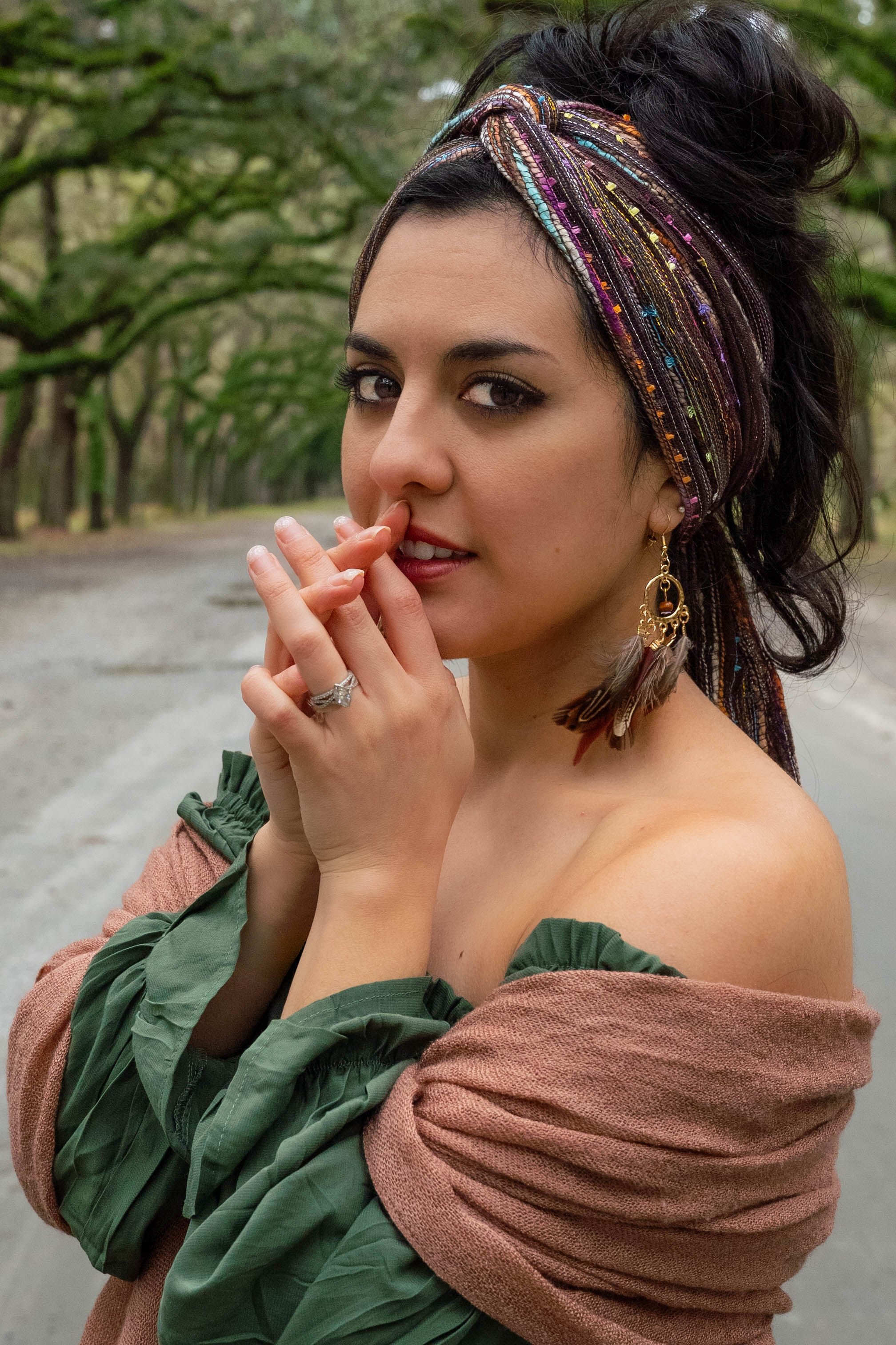 A woman with dark hair, wearing a colorful headscarf, standing outdoors surrounded by green trees, with her finger near her nose, looking at the camera.