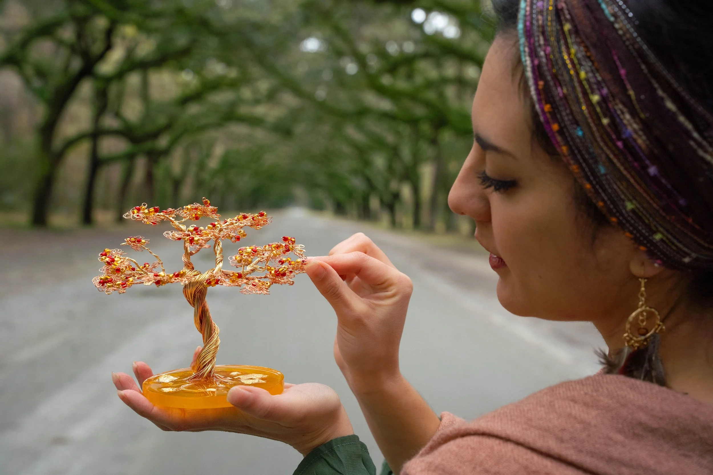 A woman with a colorful headscarf holding a decorative wire tree with red beads in her hand, on a tree-lined road.