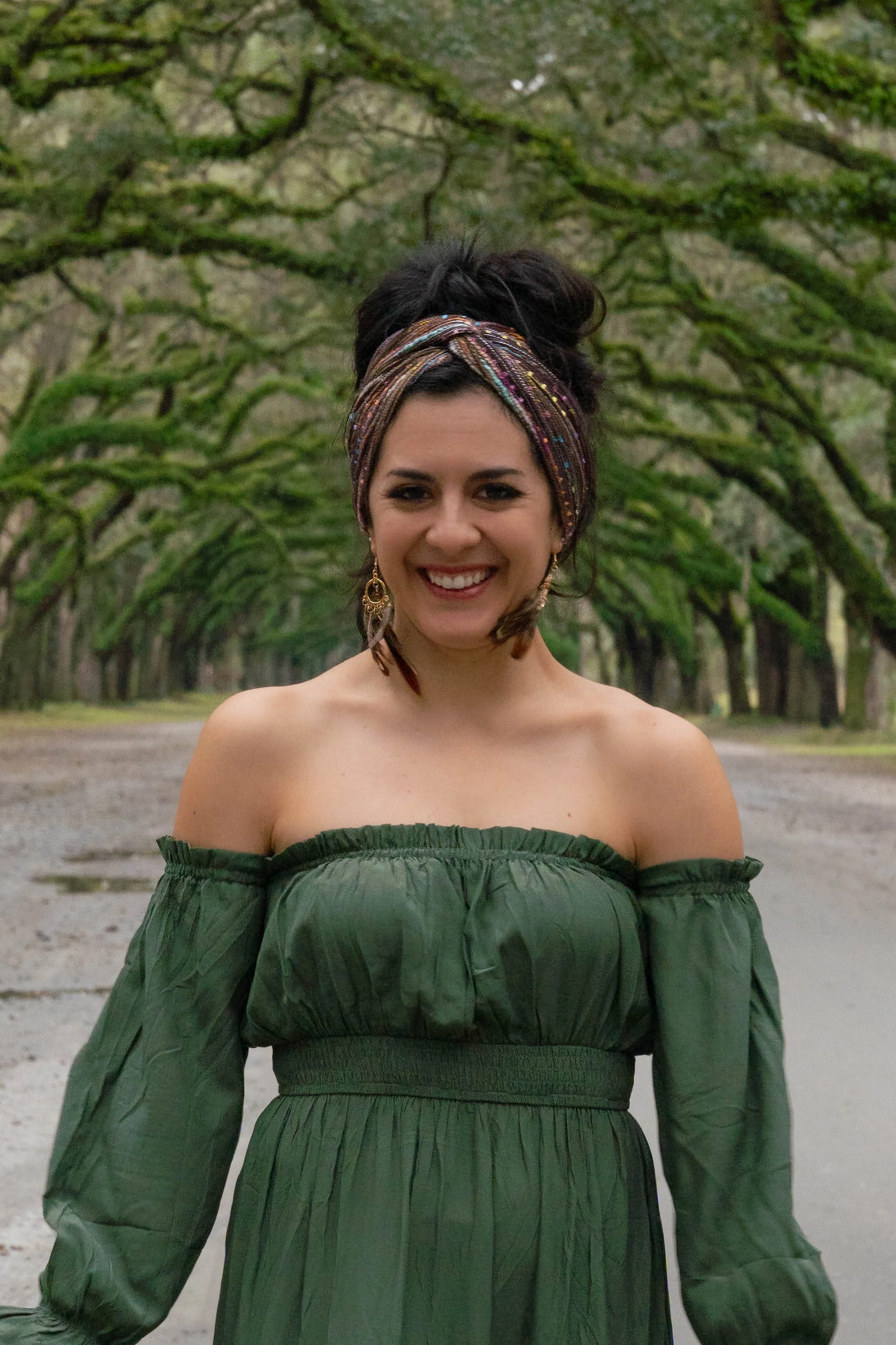 A woman smiling outdoors, standing on a tree-lined path with green moss-covered branches overhead, wearing an off-the-shoulder green dress and a colorful headscarf.
