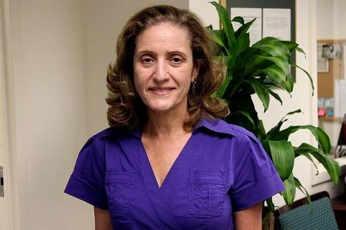 A woman with brown, curly hair wearing a purple shirt, standing indoors with a large green plant behind her.