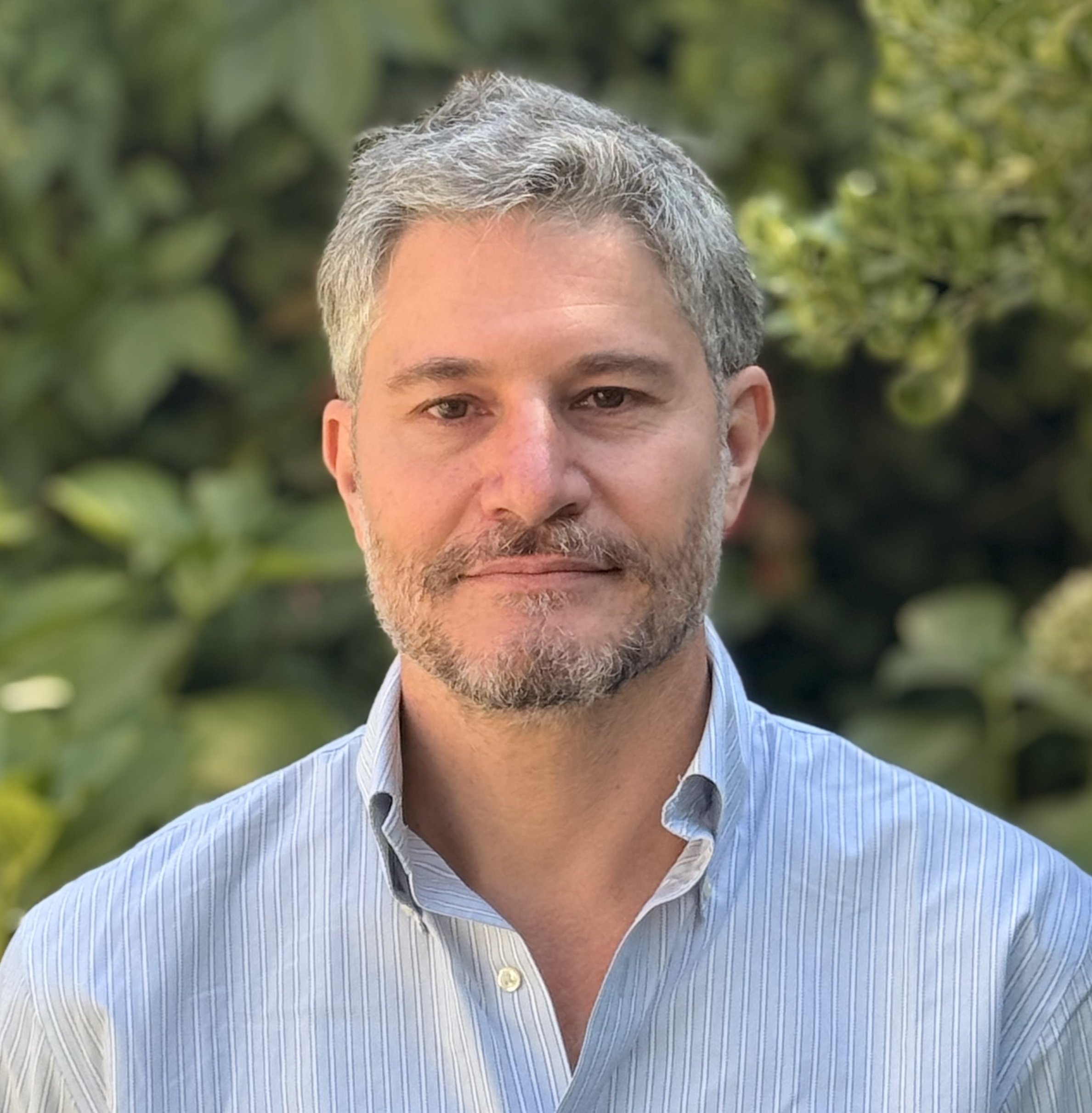 A middle-aged man with salt-and-pepper hair, a beard, and mustache, wearing a light blue striped dress shirt, standing outdoors with a blurred green leafy background.