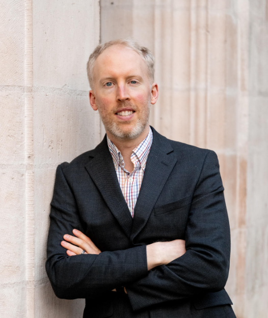 A man with light hair and beard wearing a dark suit and checkered shirt, crossing his arms and smiling, standing against a stone wall.