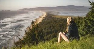 A woman sitting on a grassy hill overlooking the ocean coast with waves crashing against the shoreline and distant hills in the background.