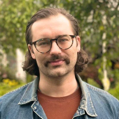 A man with glasses and a mustache posing outdoors with trees and plants in the background.