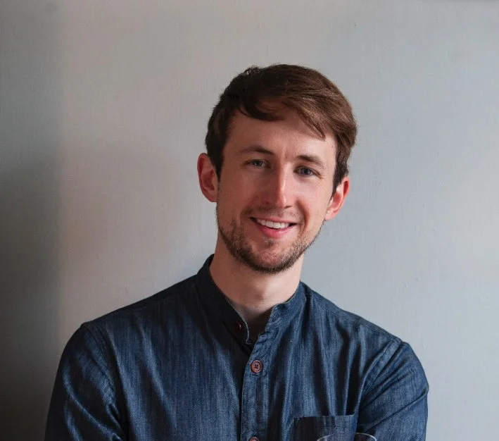 A young man with brown hair and a beard smiling, wearing a dark blue button-up shirt, standing against a plain light gray wall.