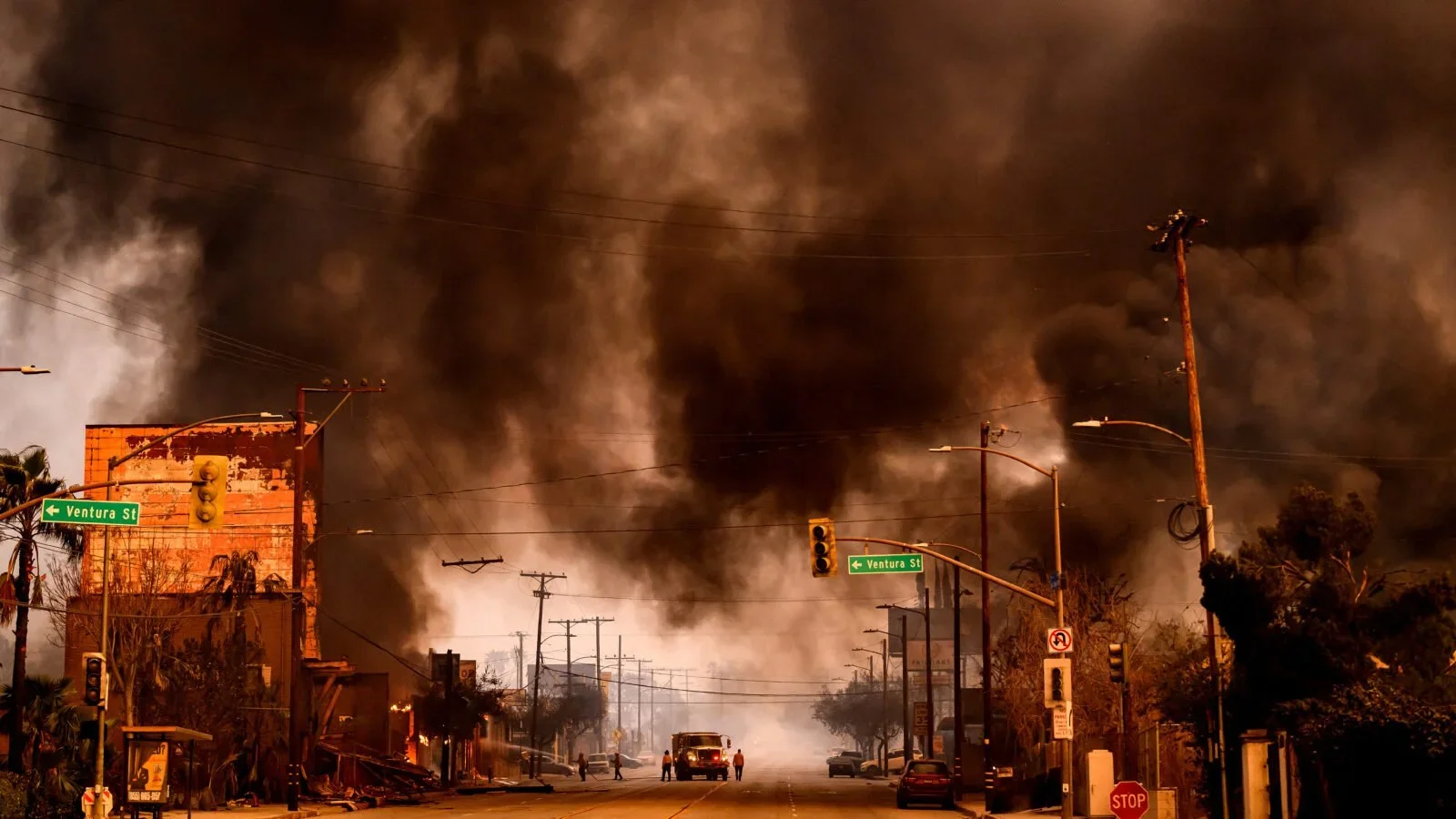 A street scene with multiple power lines and streetlights, as well as stop and traffic lights, under a dark and smoky sky caused by a large fire emitting thick black smoke in the background.
