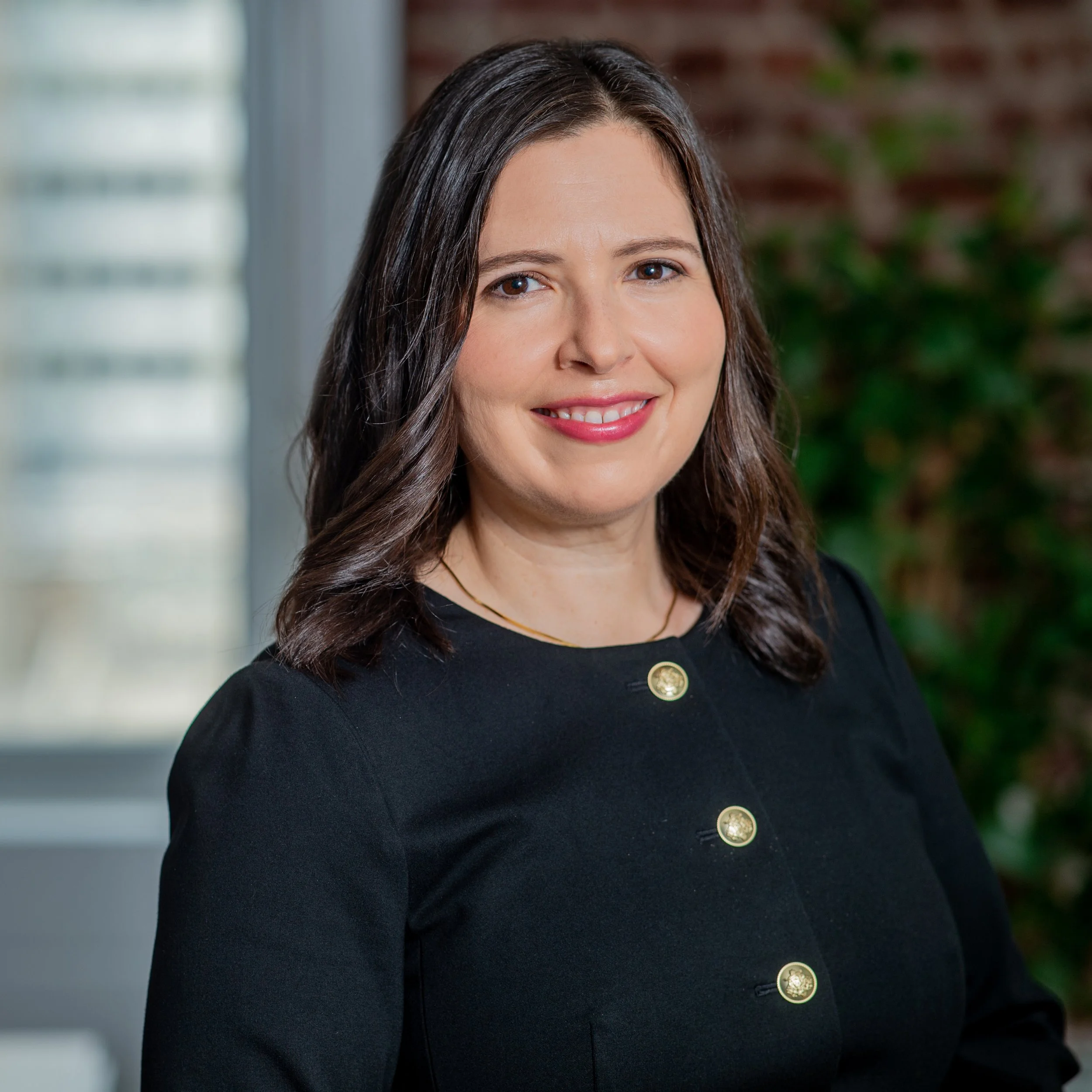 A woman with shoulder-length brunette hair, smiling, wearing a black blazer with gold buttons, standing indoors near a window.