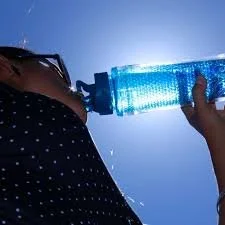 Close-up of a person pouring water from a plastic bottle onto their shoulder against a bright blue sky.