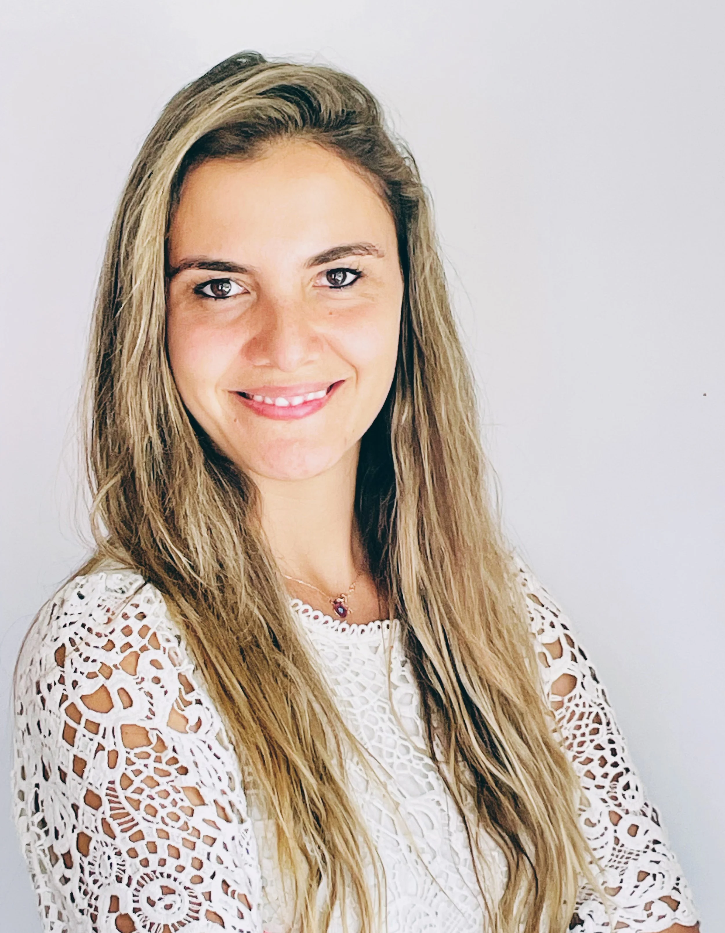 Portrait of a smiling woman with long blond hair, wearing a white embroidered lace top and a small necklace.