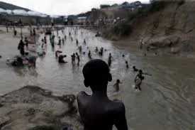 People swimming and playing in a river with a rocky shoreline and hills in the background.