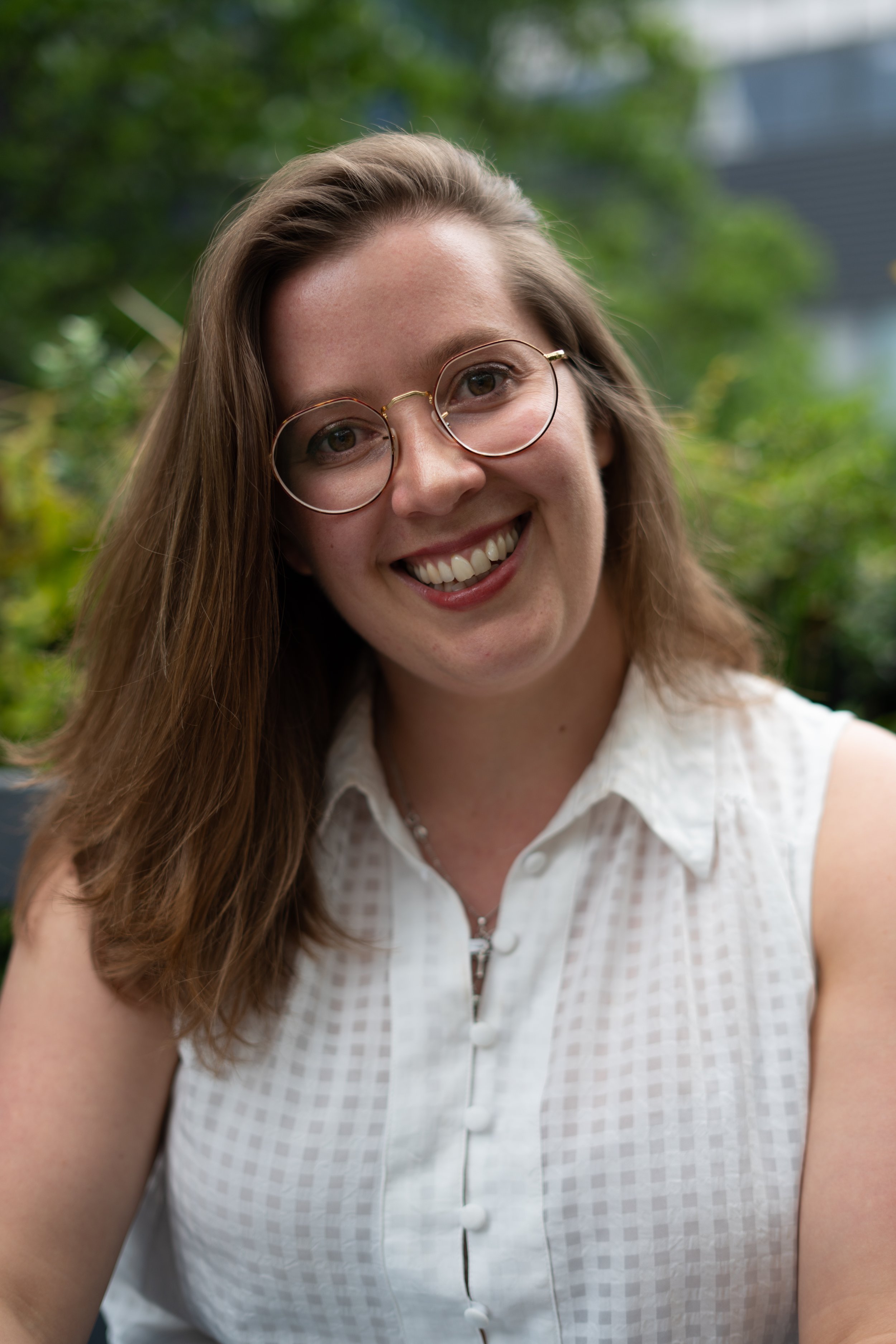 A young woman with shoulder-length brown hair, wearing glasses and a sleeveless white checkered top, smiling outdoors with blurred greenery in the background.