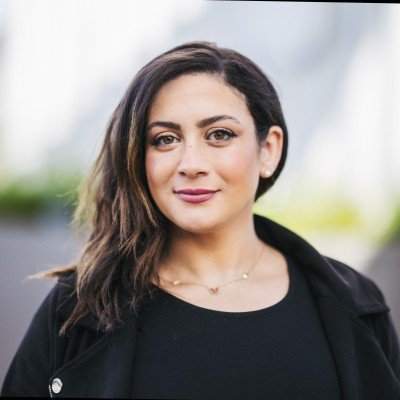 A woman with shoulder-length dark wavy hair, wearing a black top and a black jacket, standing outdoors with a blurred background.