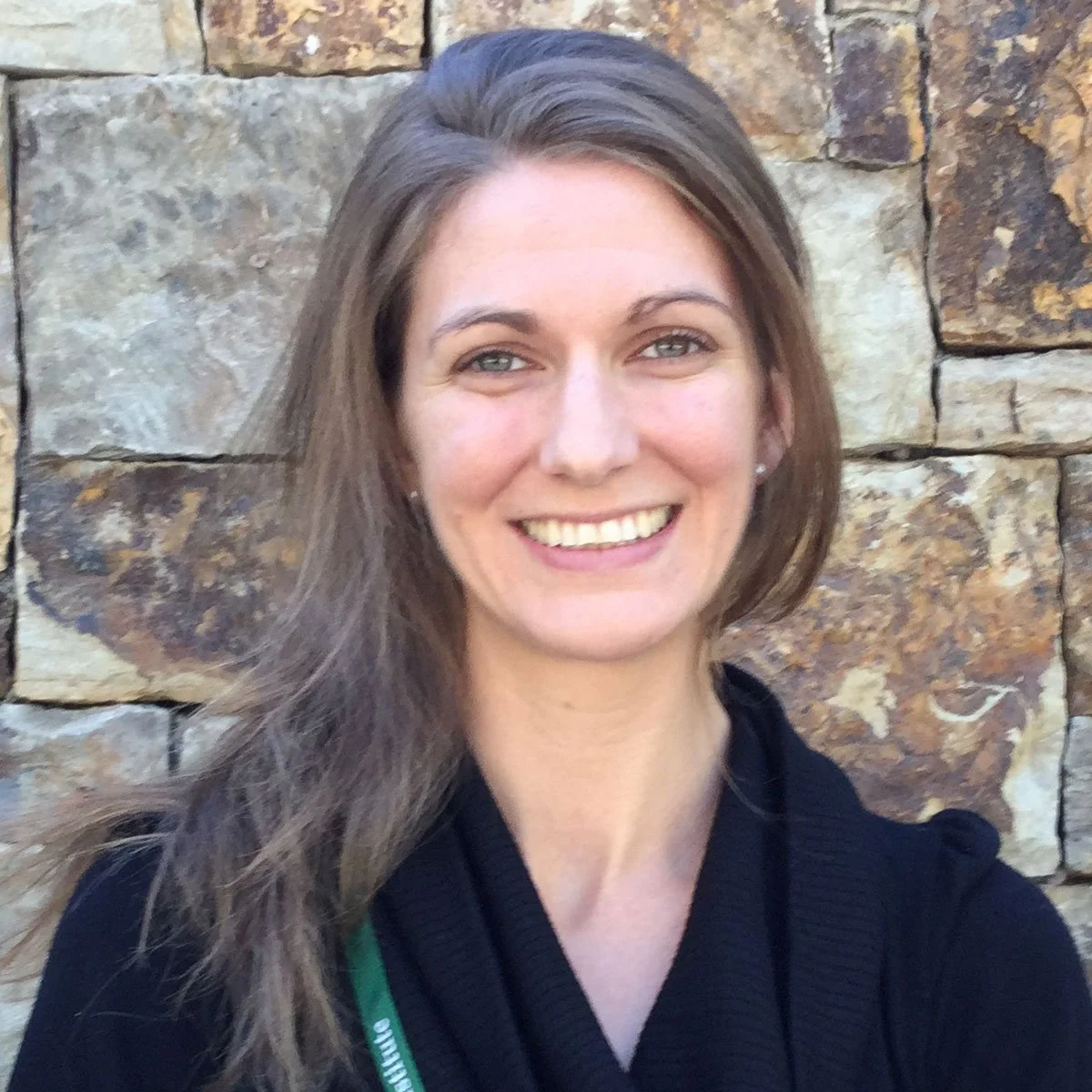 A woman with long brown hair and light-colored eyes smiling, standing in front of a stone wall.