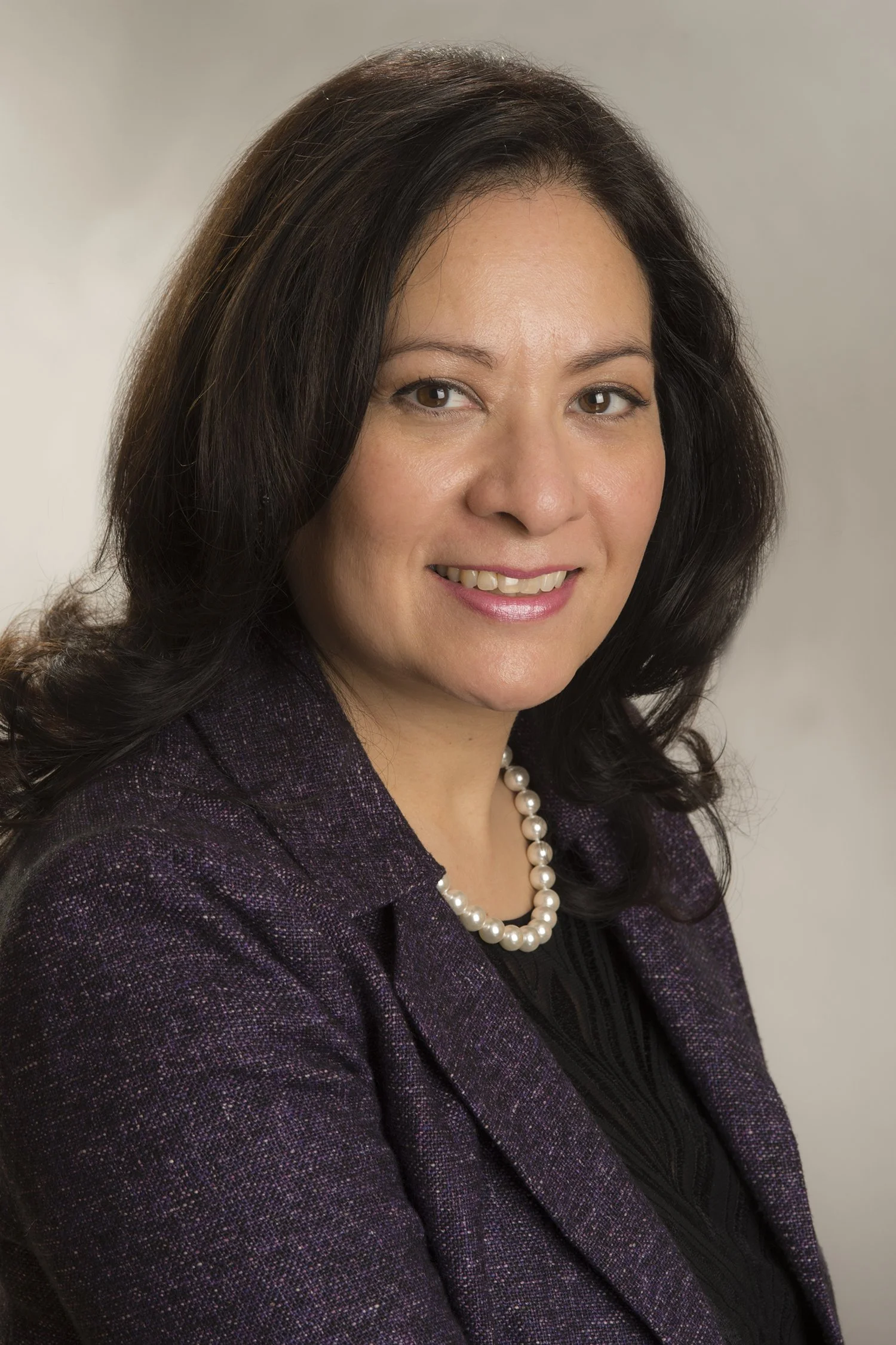 A professional woman with dark brown hair, wearing a dark purple blazer, black top, and a pearl necklace, smiling at the camera against a light, neutral background.