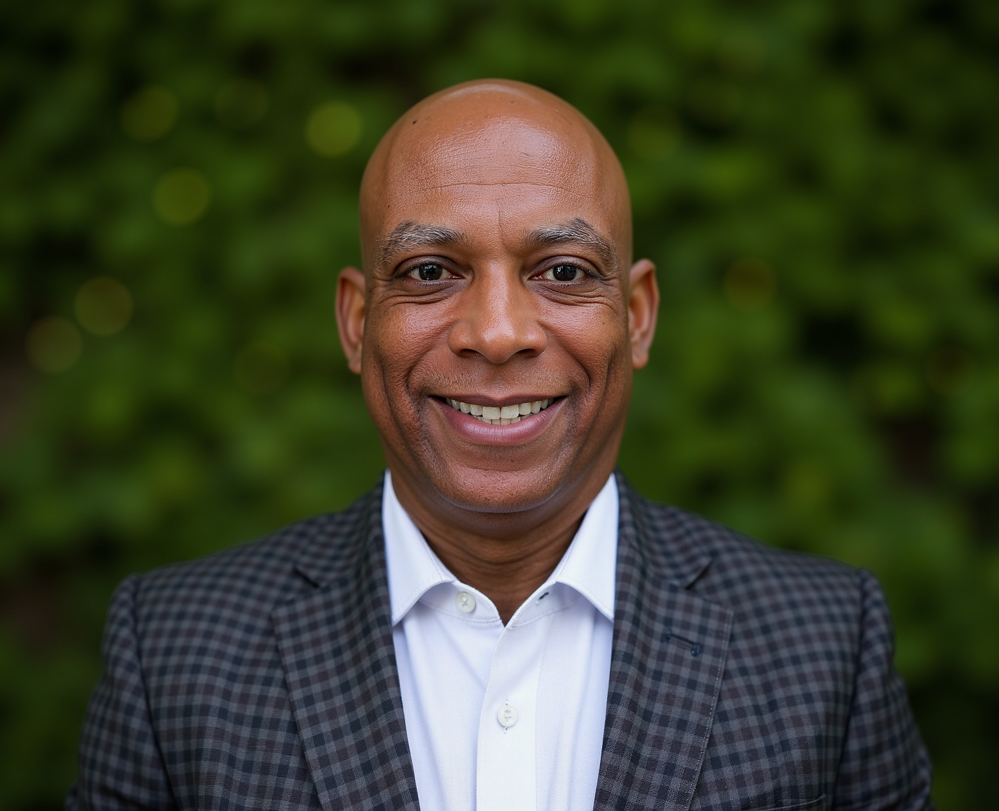Smiling man in a checkered suit and white shirt outdoors with green foliage background.
