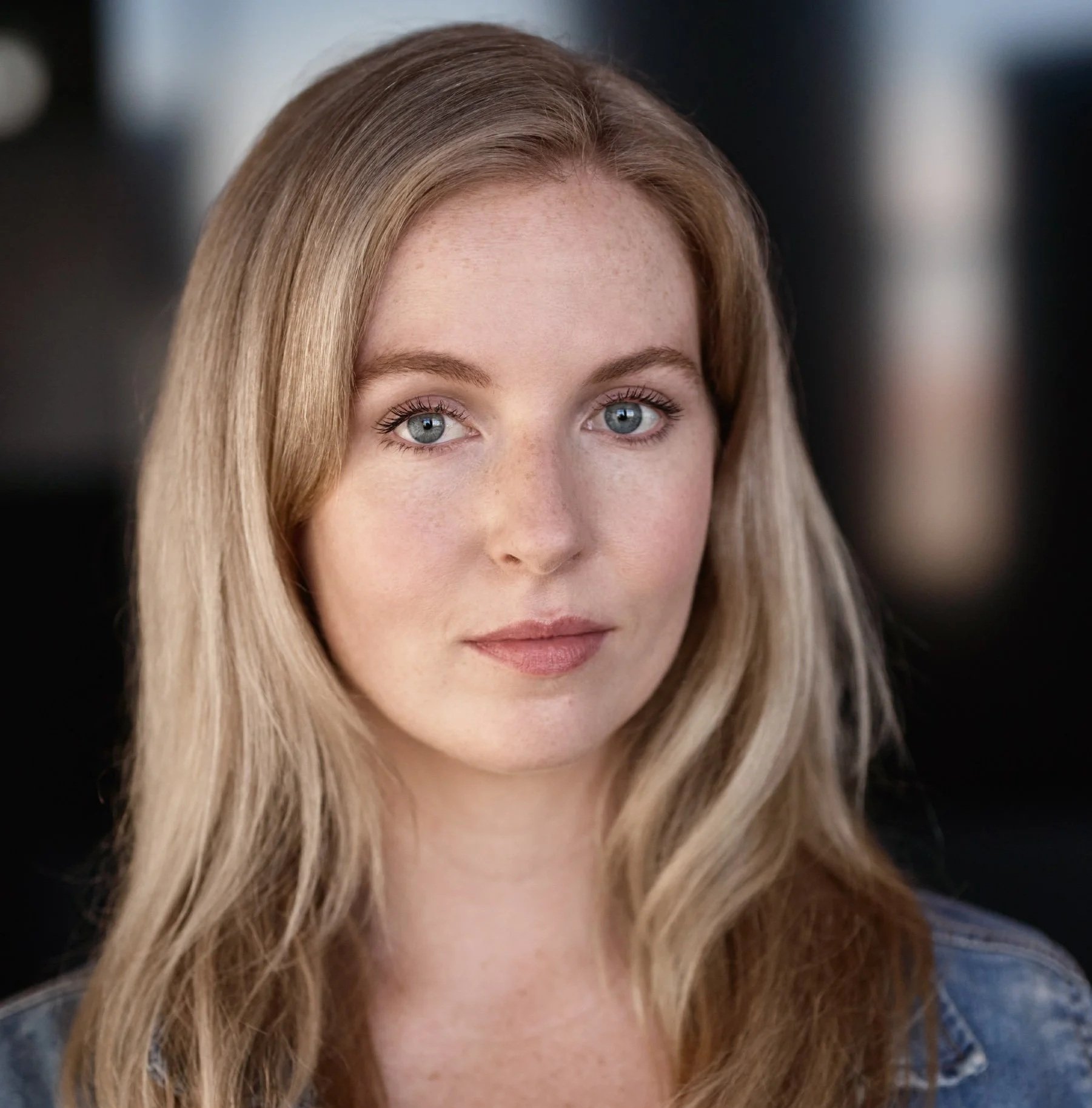 Close-up of a smiling woman with long wavy blonde hair and blue eyes against a dark background.